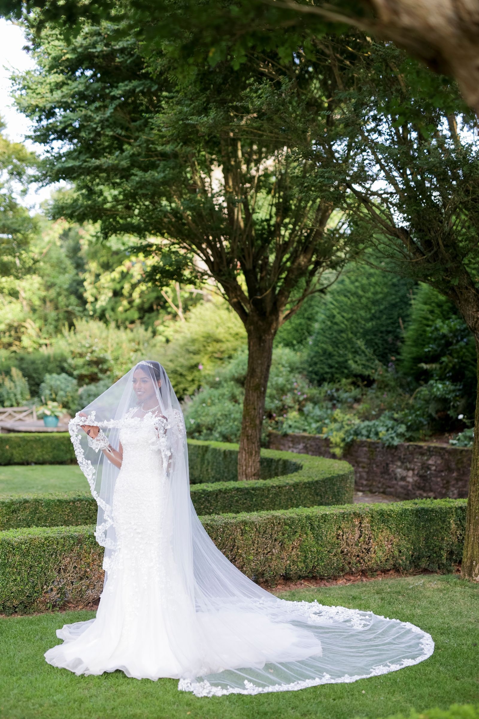 Bride alone on the lawn between clipped hedges, her cathedral veil spread out