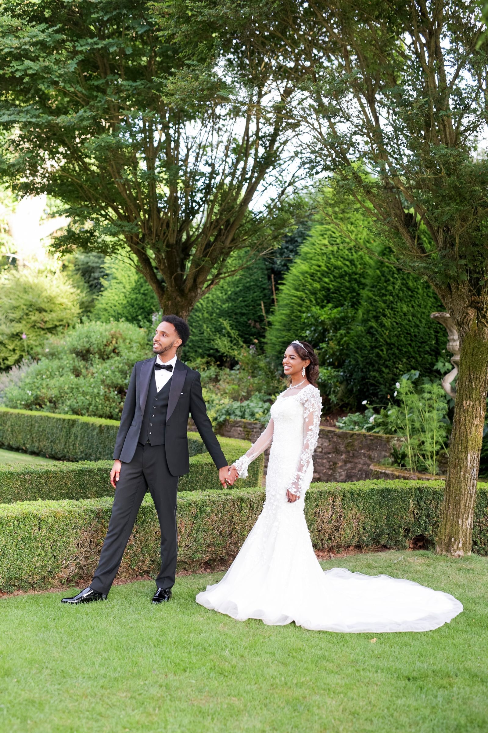 Bride and groom holding hands on the lawn, hedges and trees around them