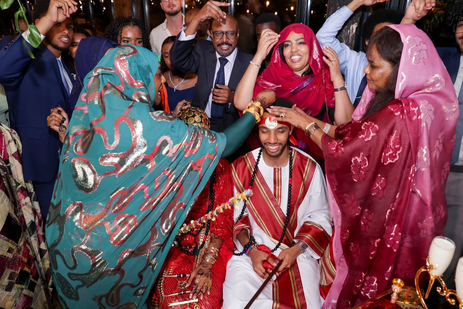 The couple seated in red and white traditional dress as family bless them, guests crowding round