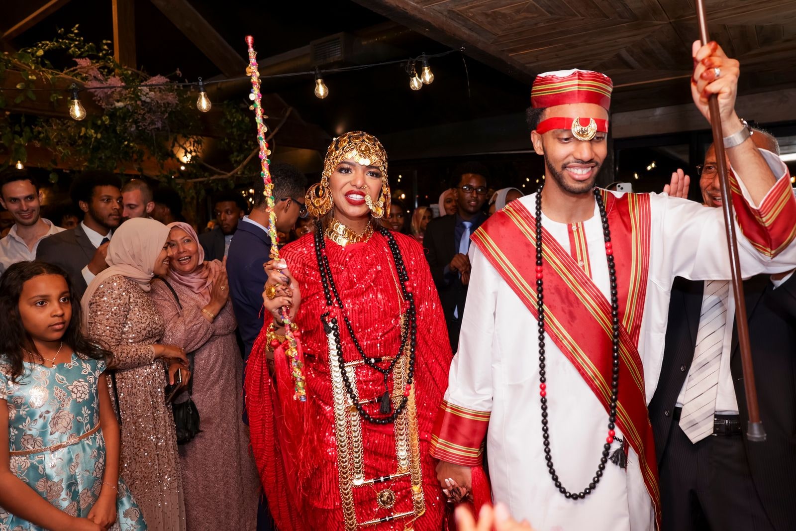 Bride and groom in traditional red and white, she holding a decorated staff, guests around them