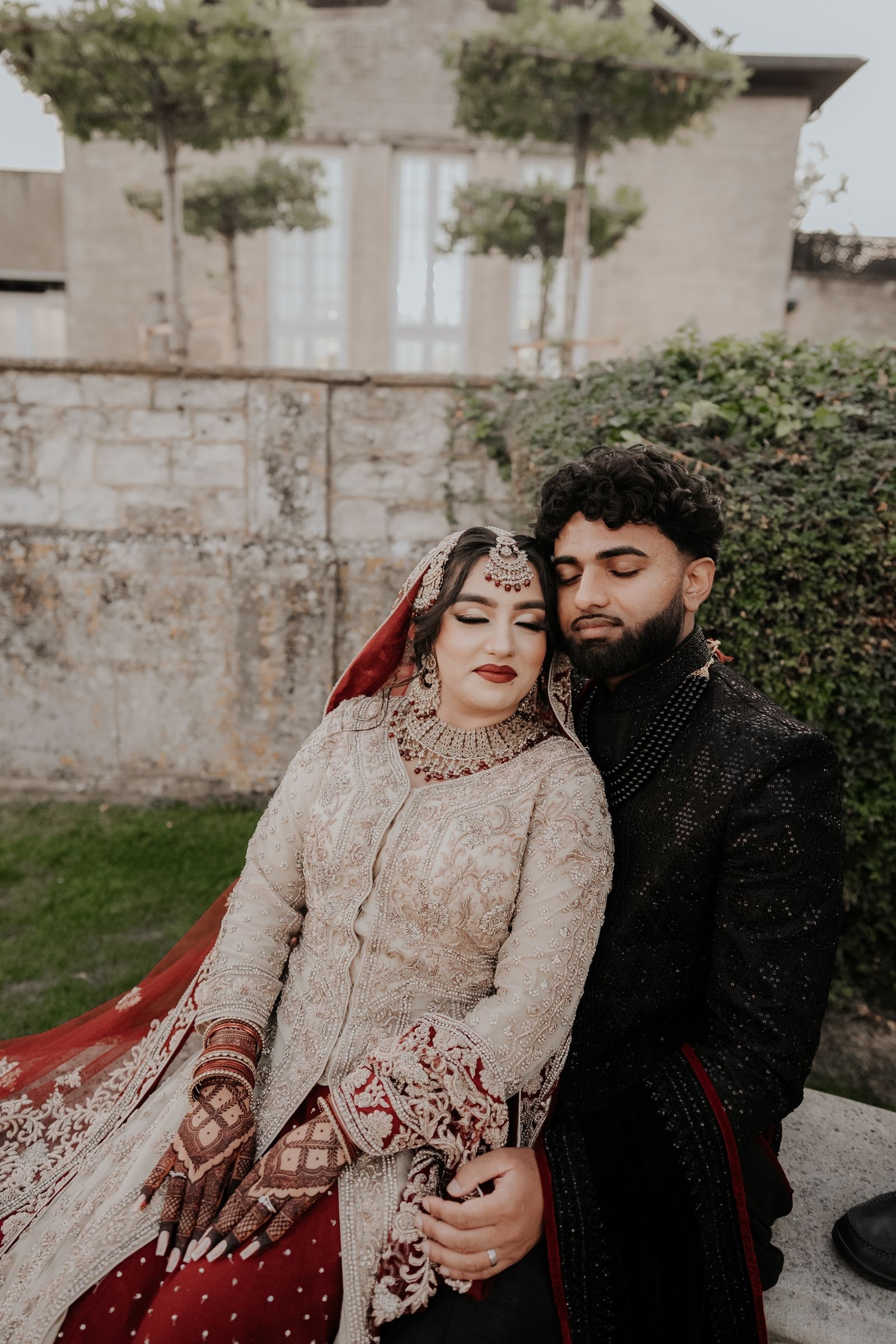 Bride in ivory and red with her groom in a black sherwani, seated by a stone wall
