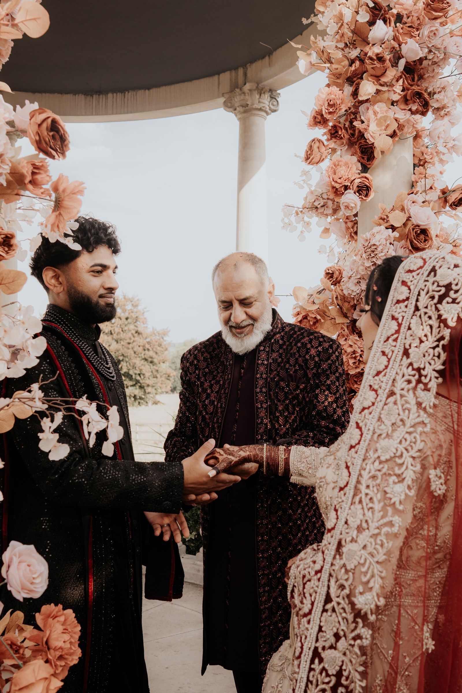 The couple's hands joined by an elder during the ceremony, before a wall of peach roses