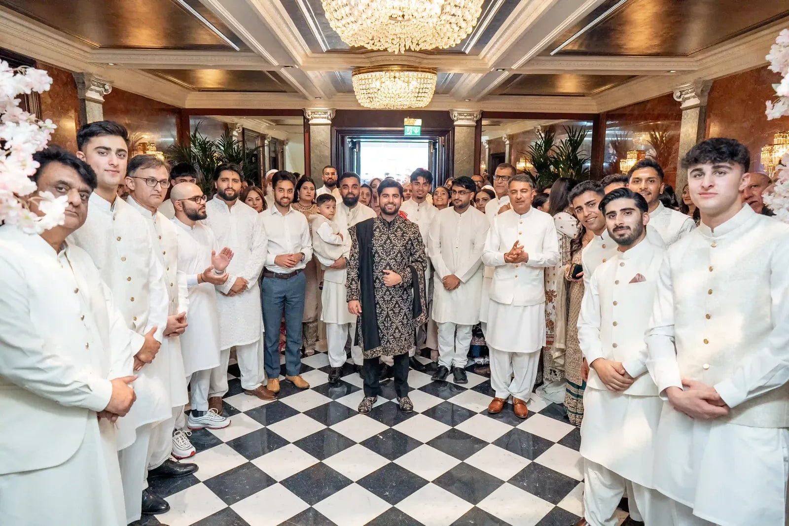 Groom standing alone on the chequered floor, his friends in ivory in a ring around him