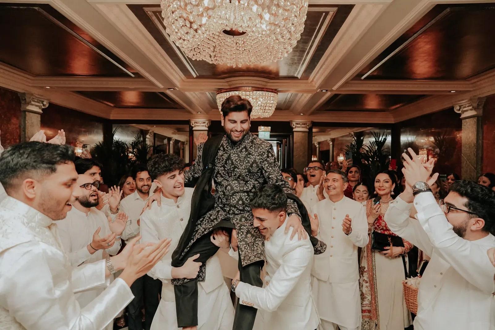 Groom carried on shoulders beneath the chandeliers as his friends applaud
