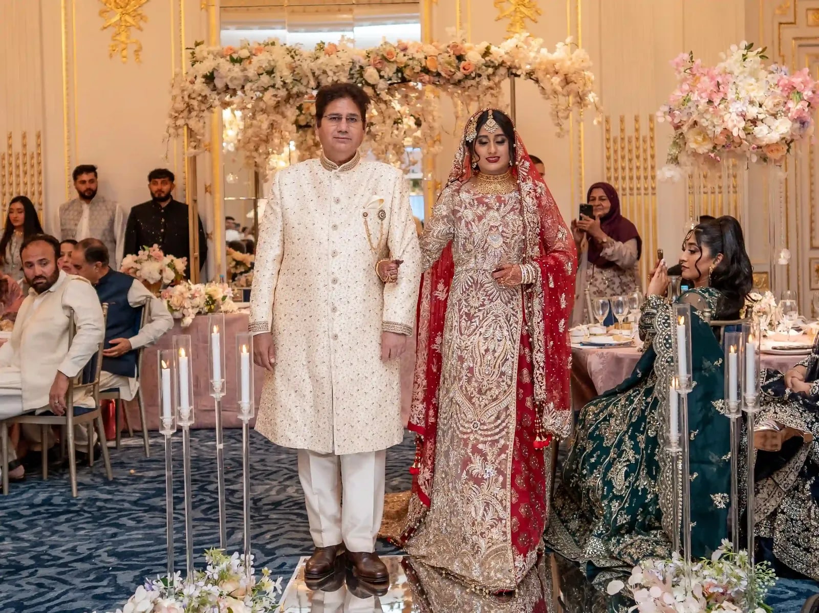 Bride and her father standing at the flower arch, guests seated at the tables