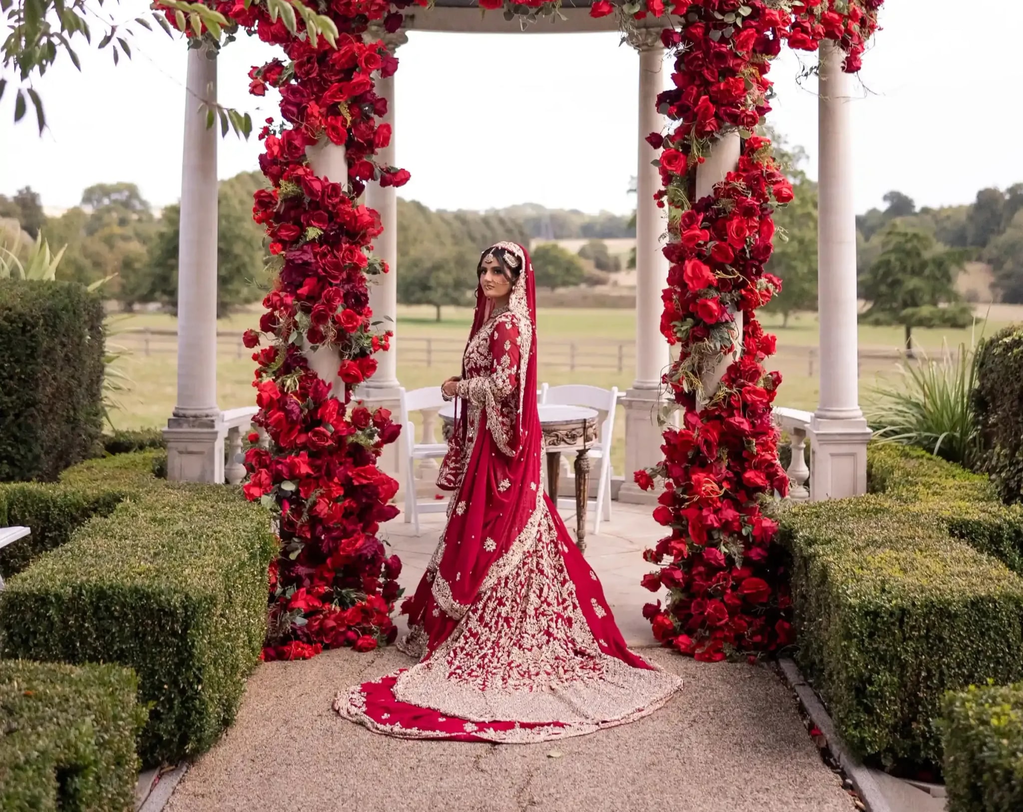 Bride standing under the red rose arch, the fields beyond stretching away