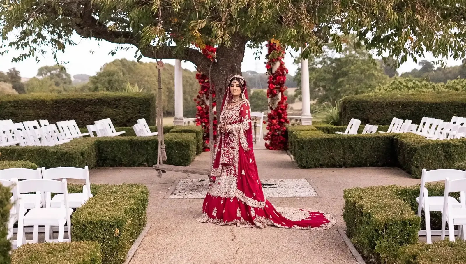 Bride small in the garden under a wide tree, rows of white chairs either side