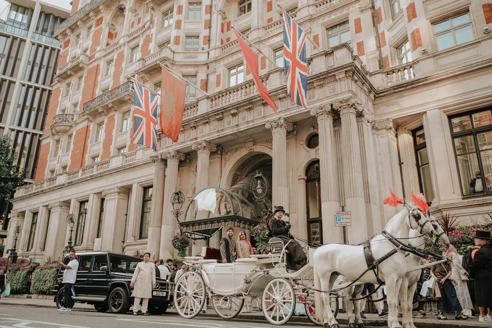 The couple in a white horse-drawn carriage outside the hotel, guests looking on