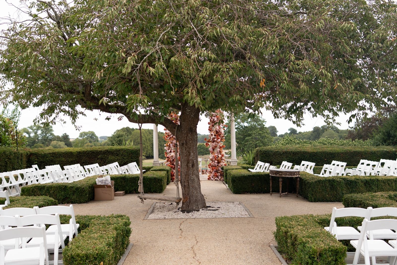 The outdoor ceremony set out under a spreading tree, white chairs between clipped hedges