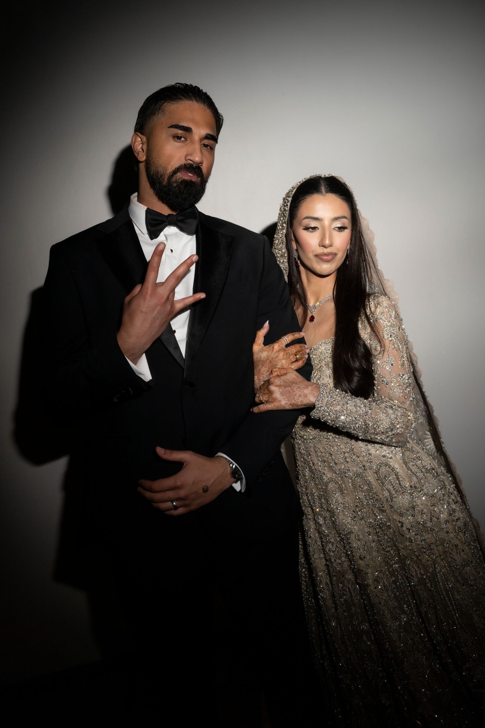 Bride and groom lit hard against a pale wall, she in gold, he in black tie