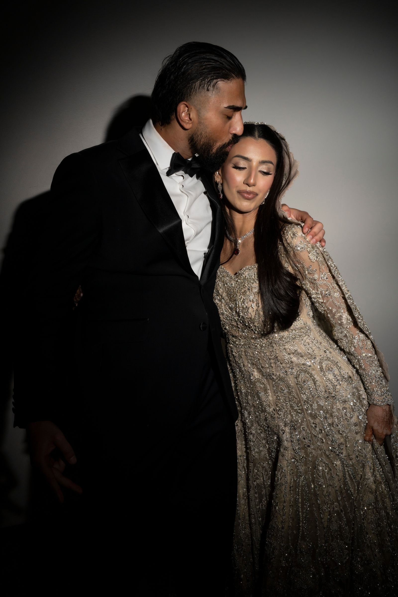 Groom leaning down to his bride's head, both lit against a dark wall