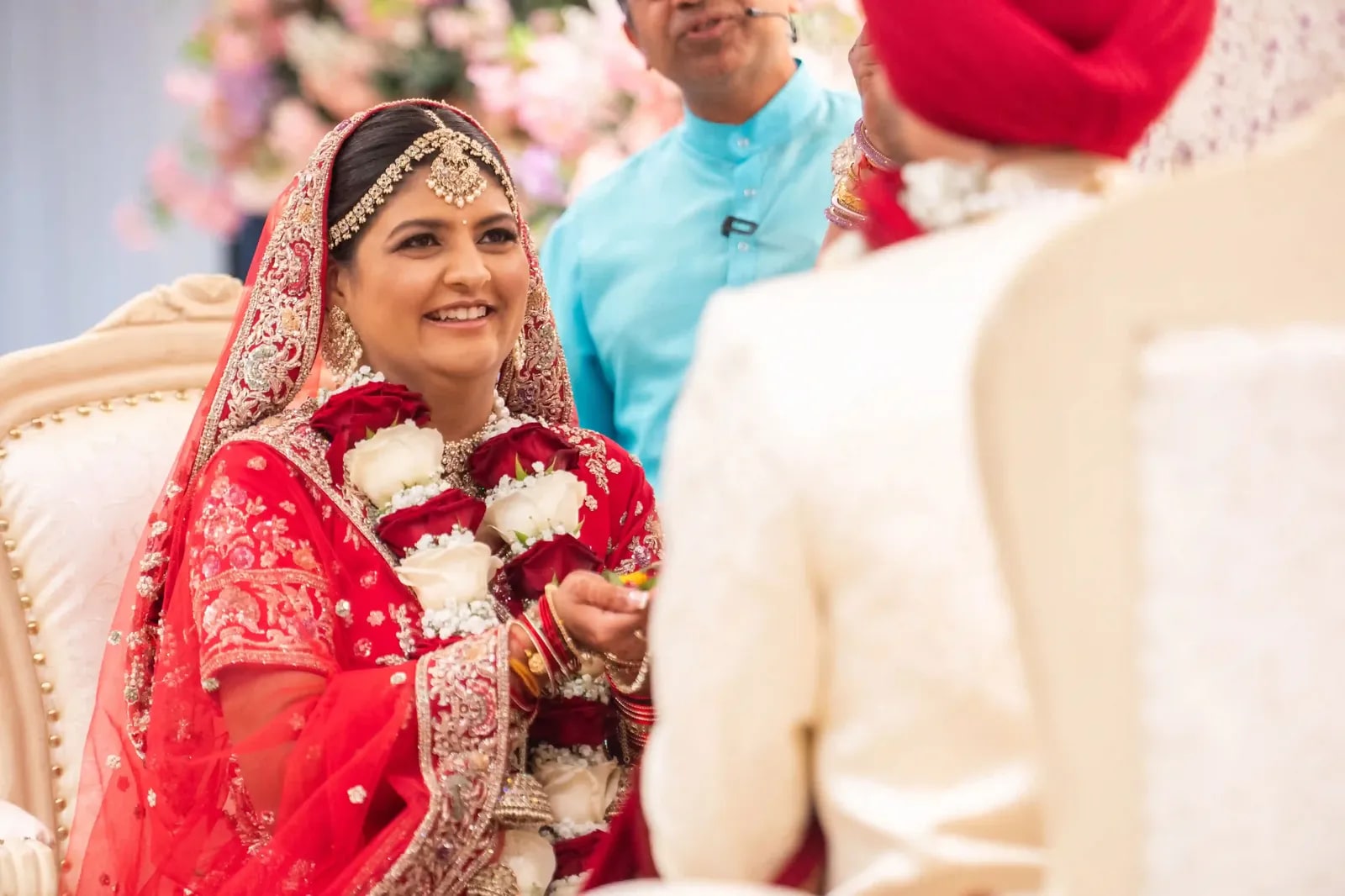 Bride smiling in a red lehenga and flower garland, her groom in the foreground