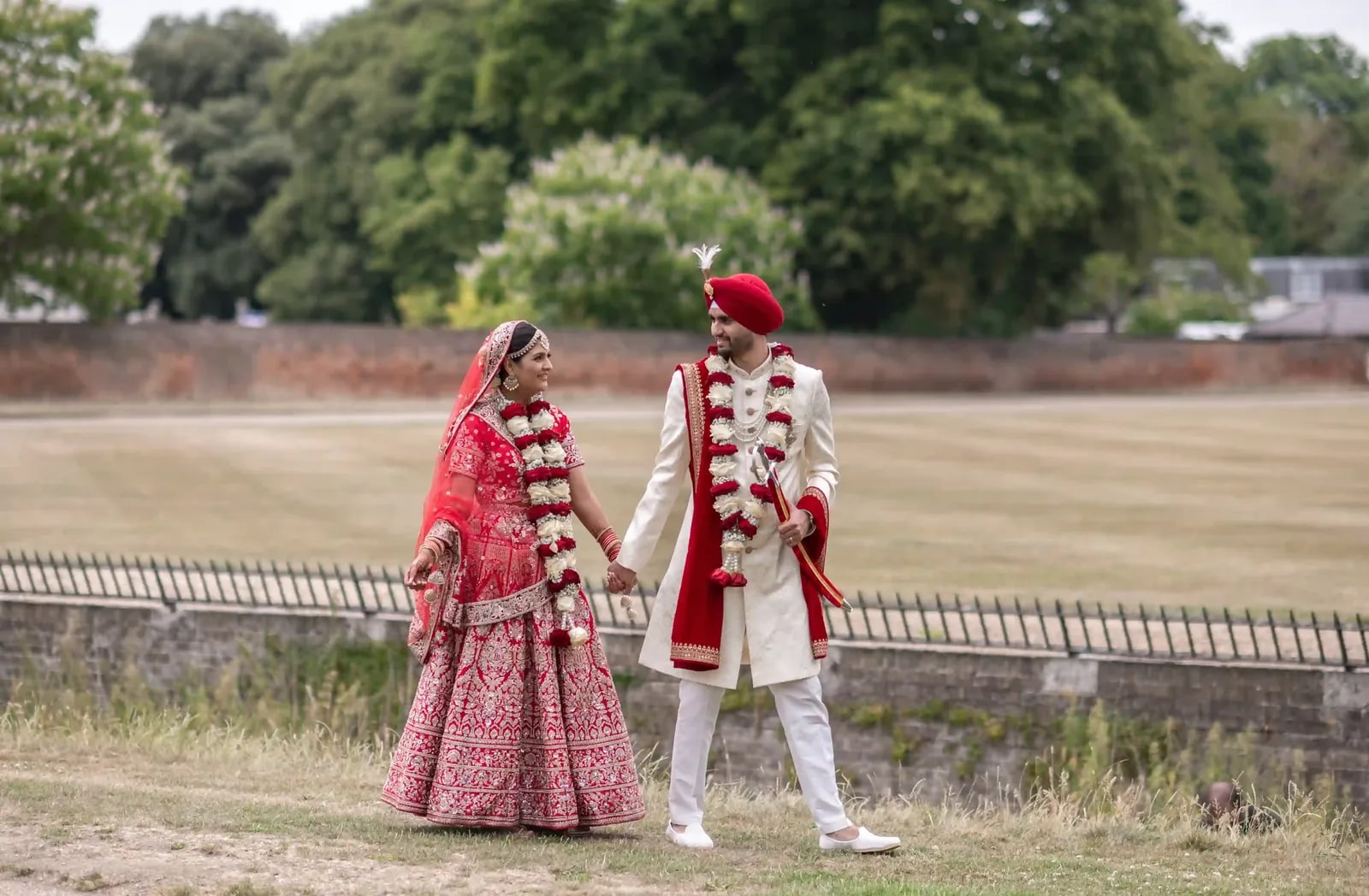 Bride and groom walking together across the grass beside a low wall