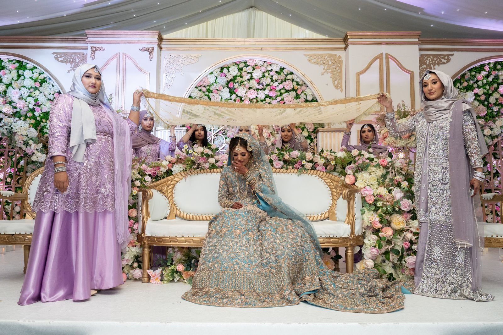 Bride seated alone on the stage sofa as her party hold a veil above her