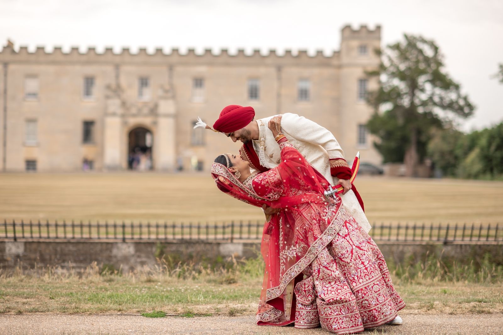 Groom in a red turban dipping his bride in a red lehenga on the lawn in front of Syon House