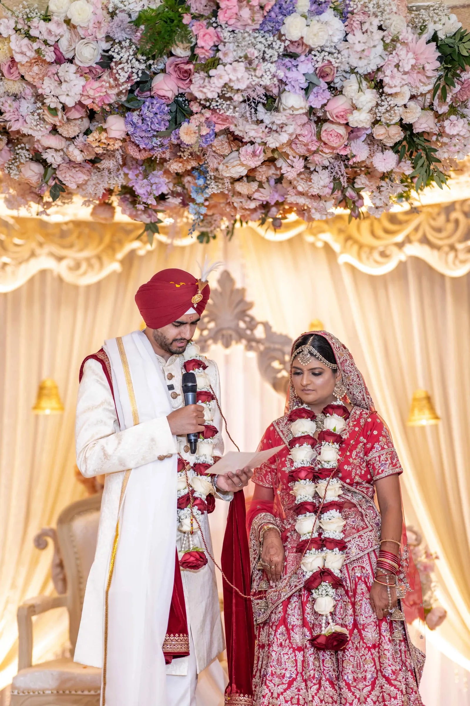 Bride and groom garlanded at the mandap, he reading from a card into a microphone