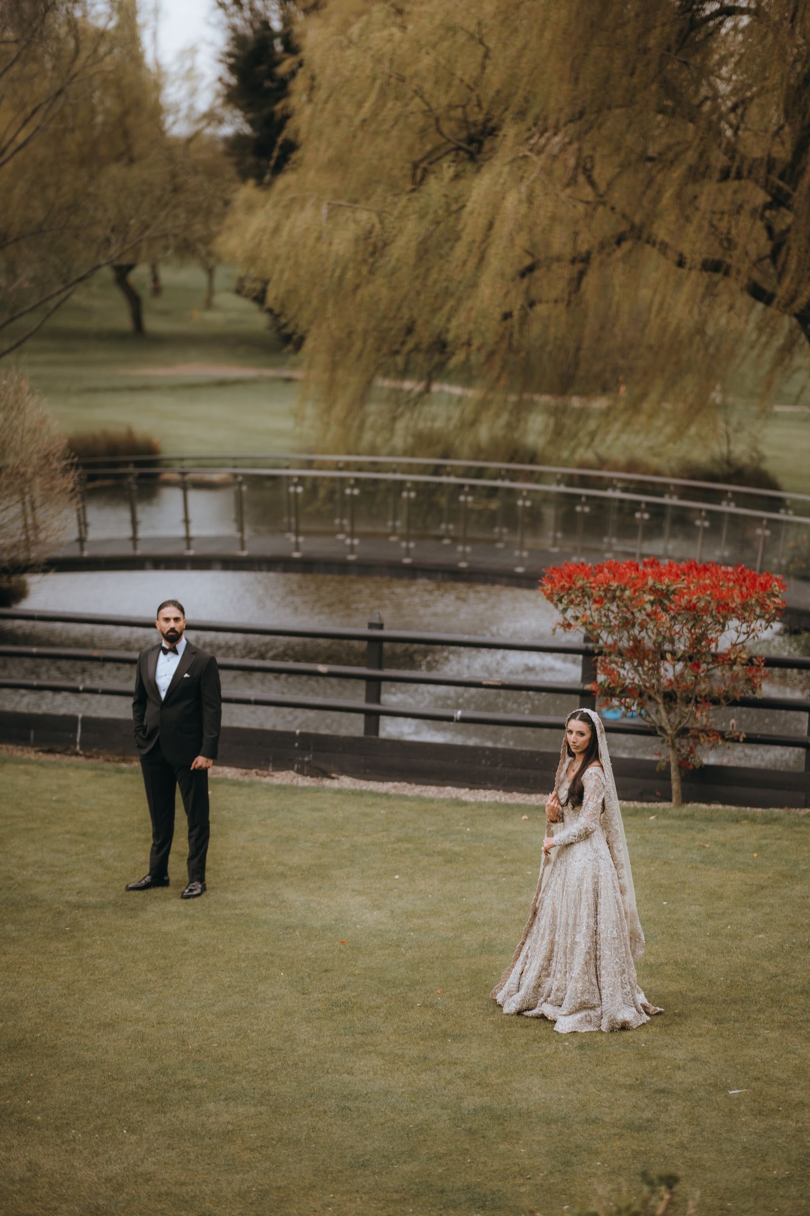 Bride and groom standing apart on the lawn, willows and the lake behind them