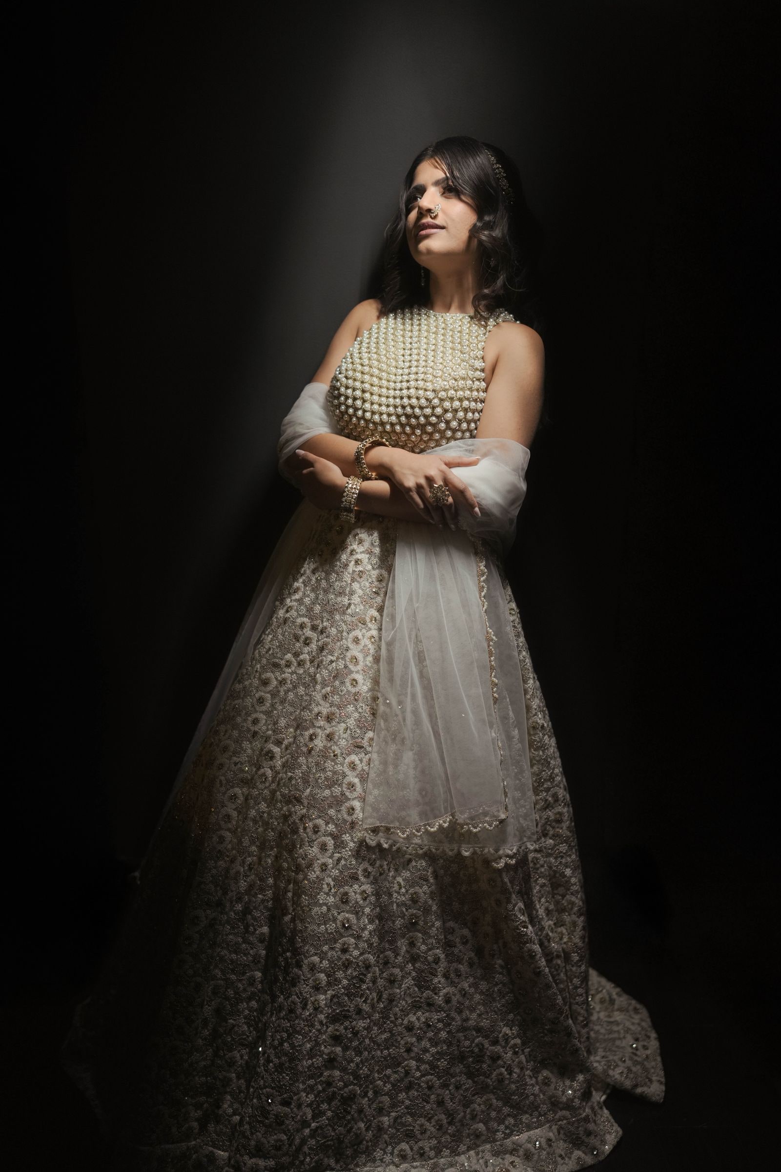 Bride standing alone in an ivory and gold lehenga, lit against a dark background