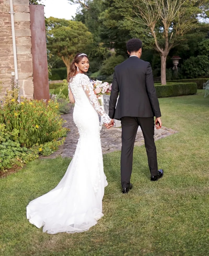 Bride and groom walking up the garden path, her lace train trailing across the grass