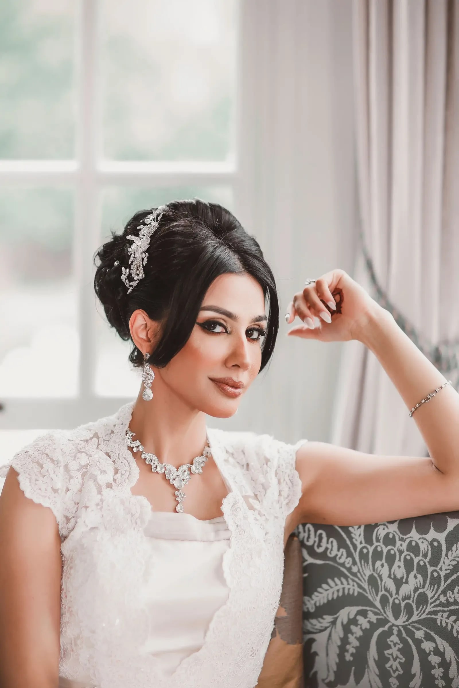 Bride seated by a window in a lace gown and diamond necklace