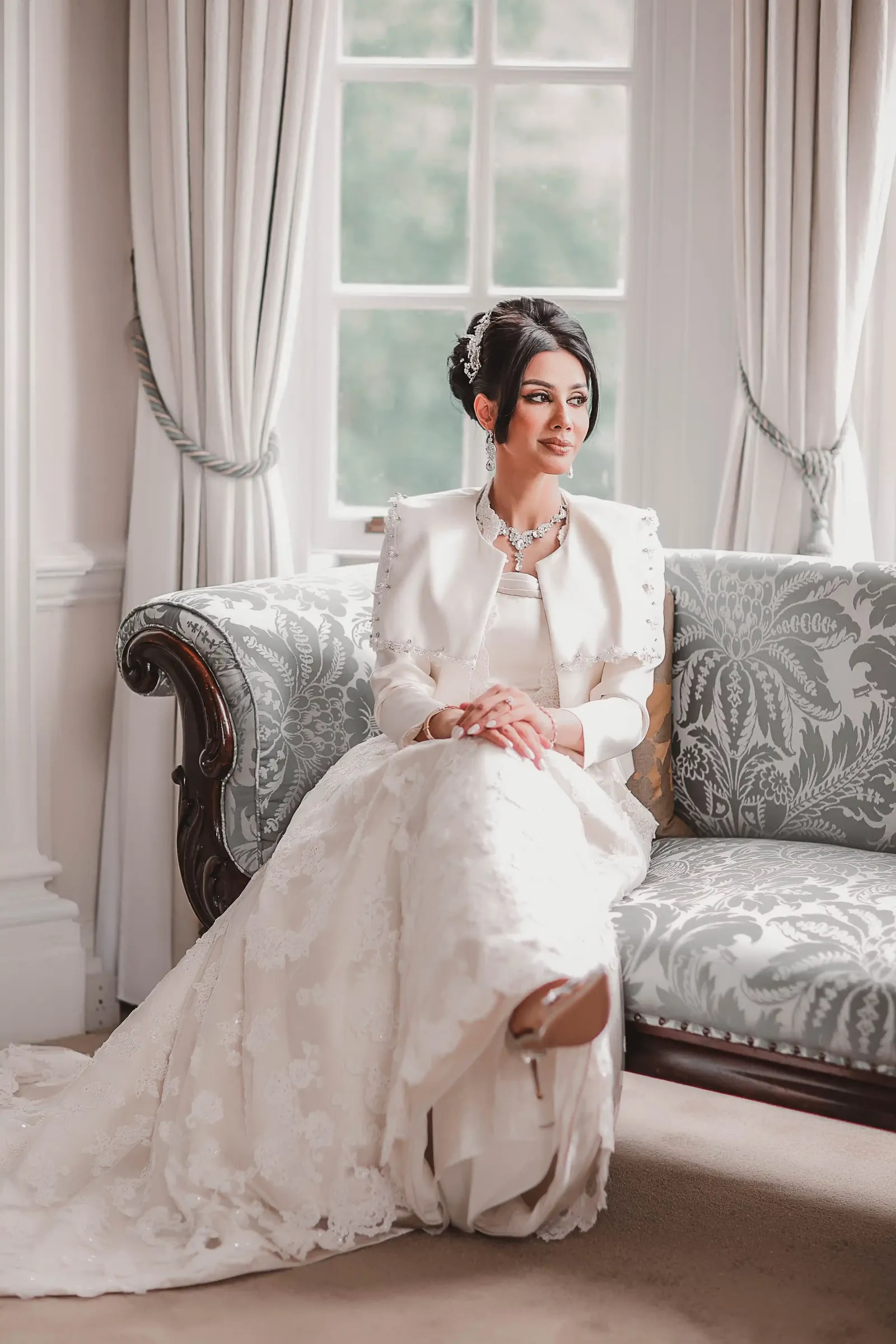 Bride seated on a chaise longue by a tall window, her lace train across the floor