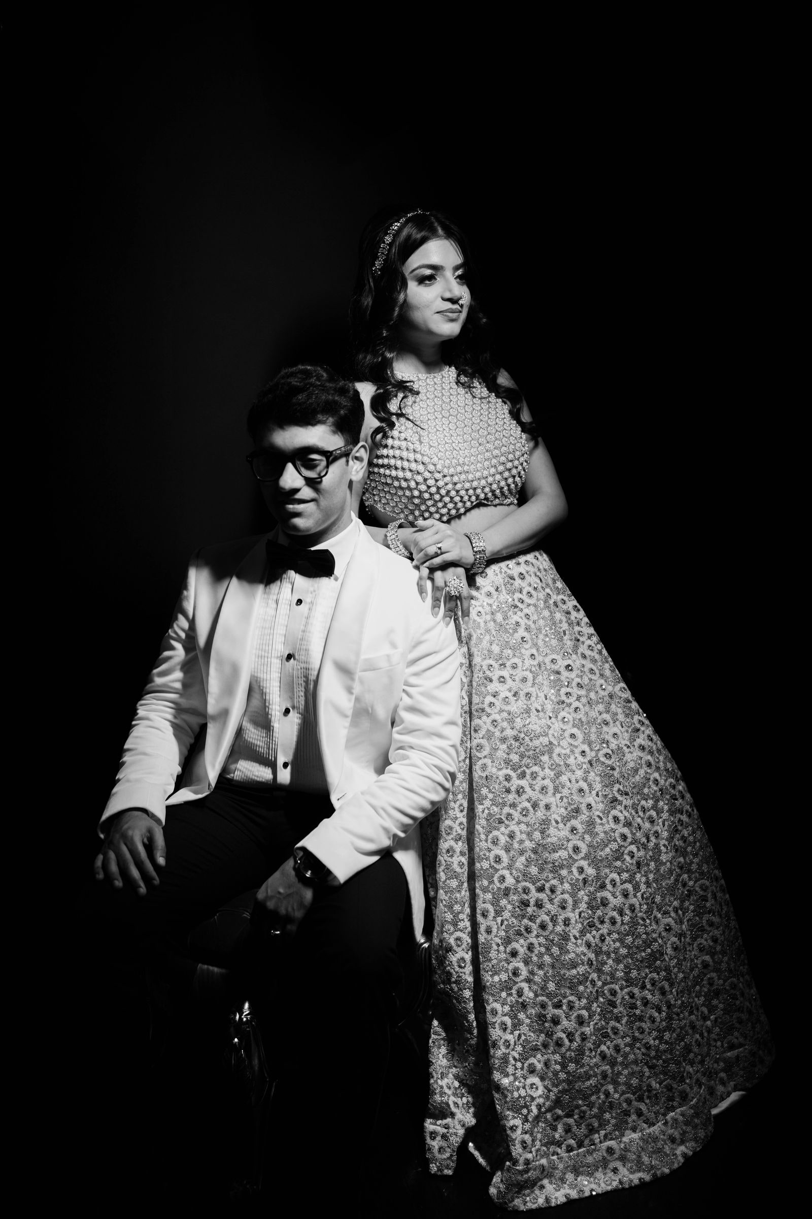 Groom seated with his bride standing behind him, both lit against black