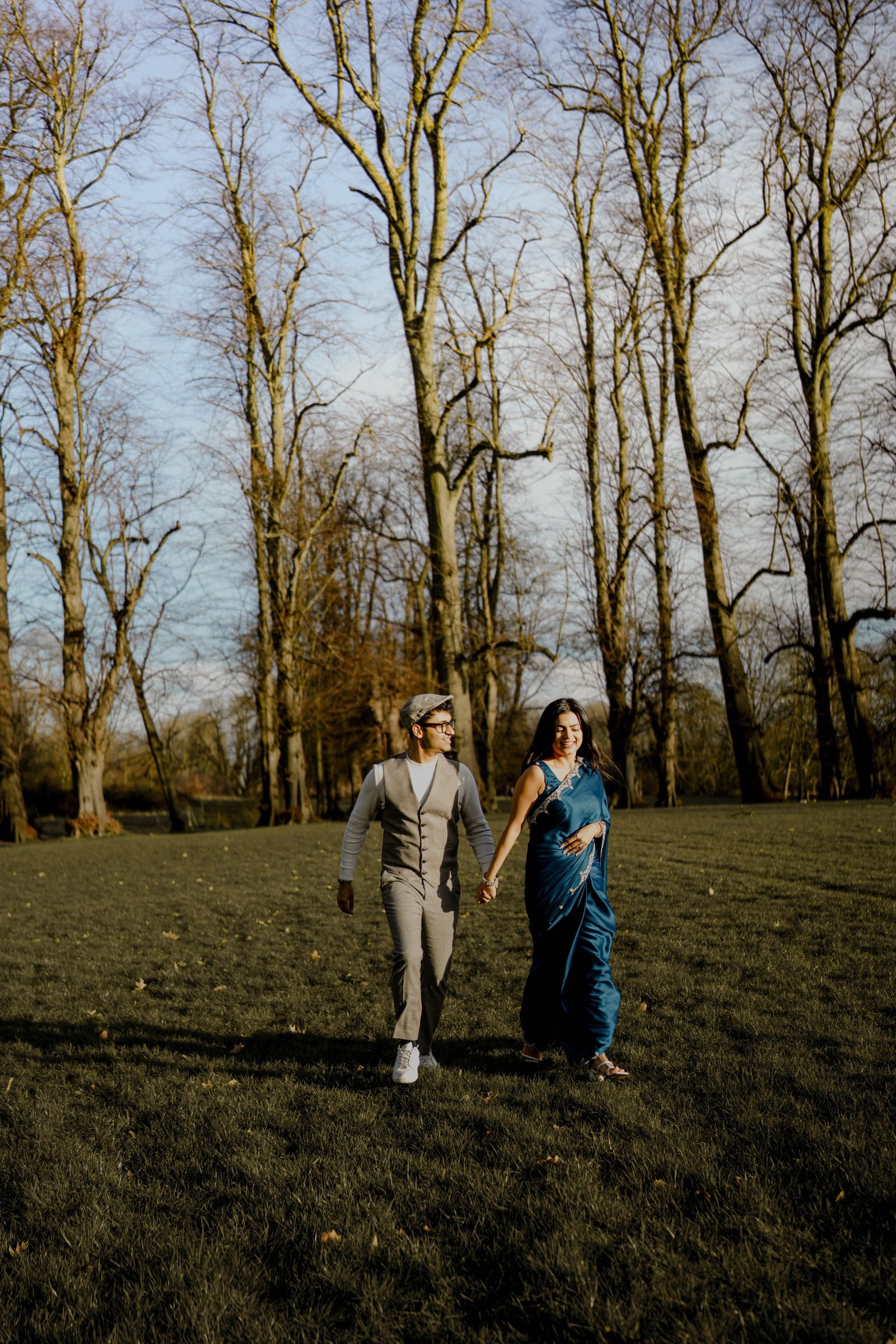 Couple walking hand in hand through bare winter trees, she in a teal sari