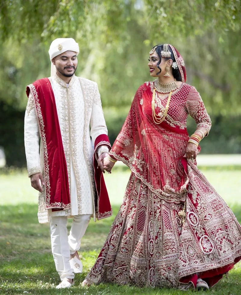 Bride and groom walking hand in hand across the grass, her red lehenga catching the light