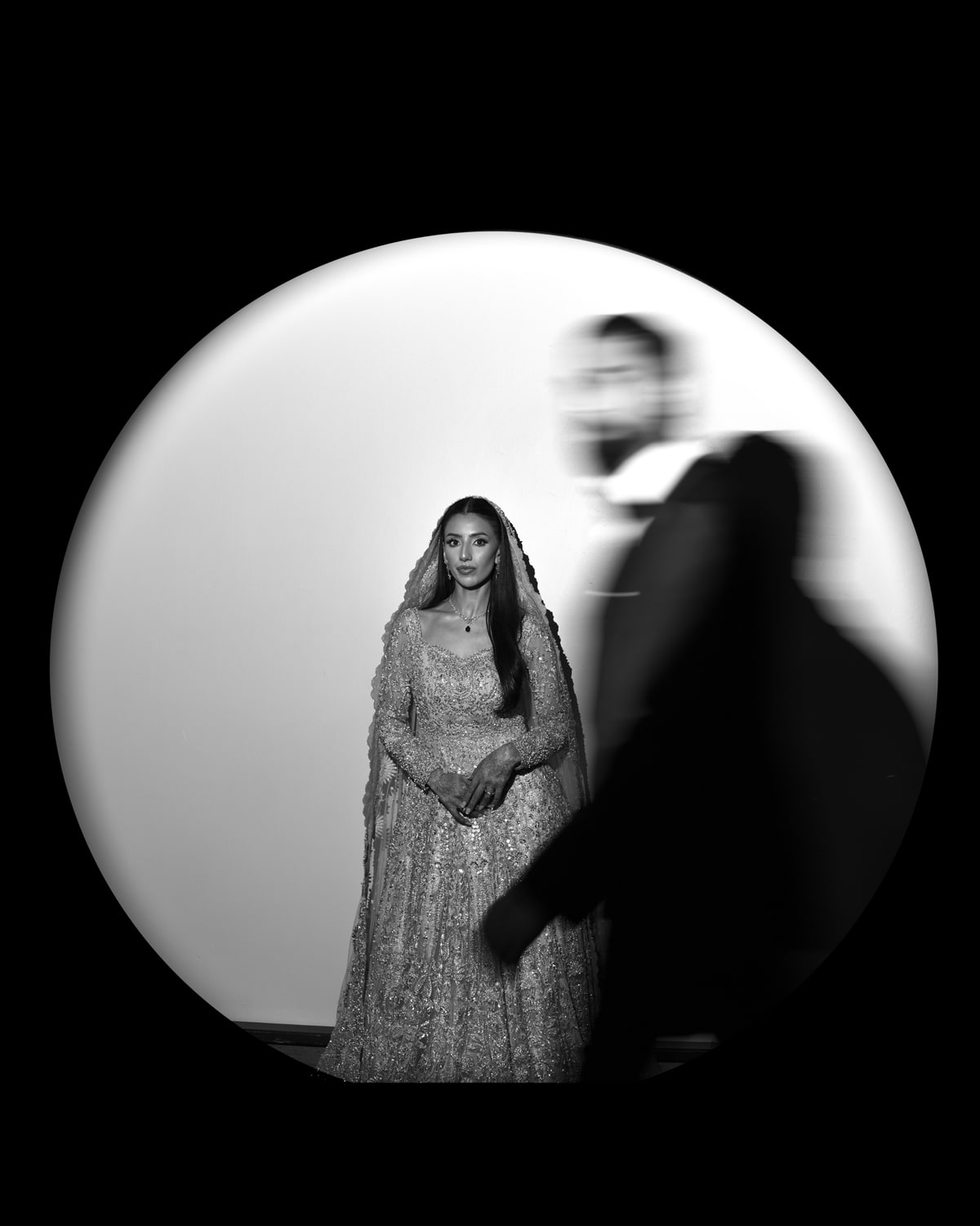 Bride standing in a circle of light with her groom passing as a blur, in black and white
