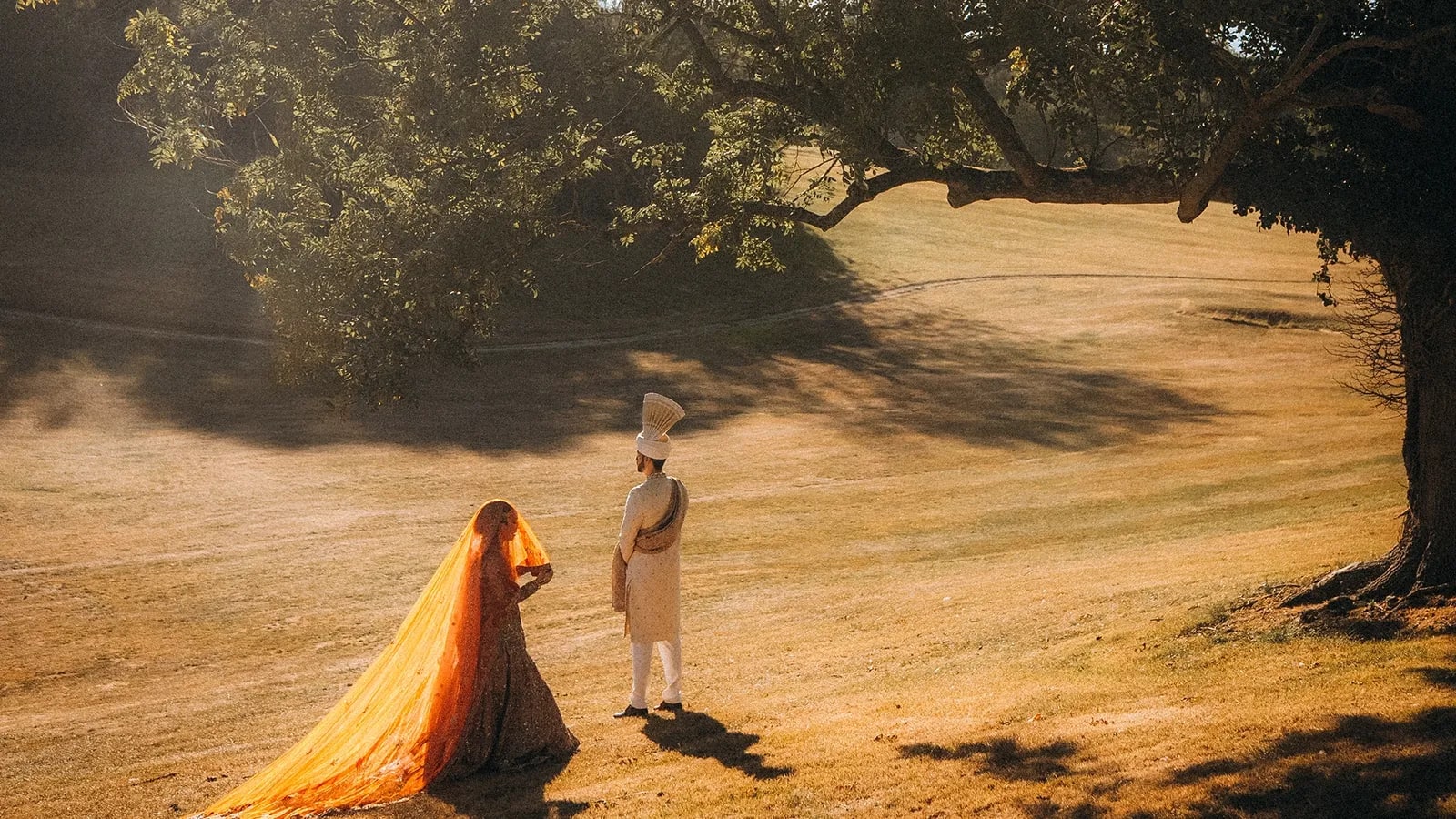 Muslim bride in a gold and orange dupatta with a long train facing her groom in a cream sherwani and turban, in a sunlit field