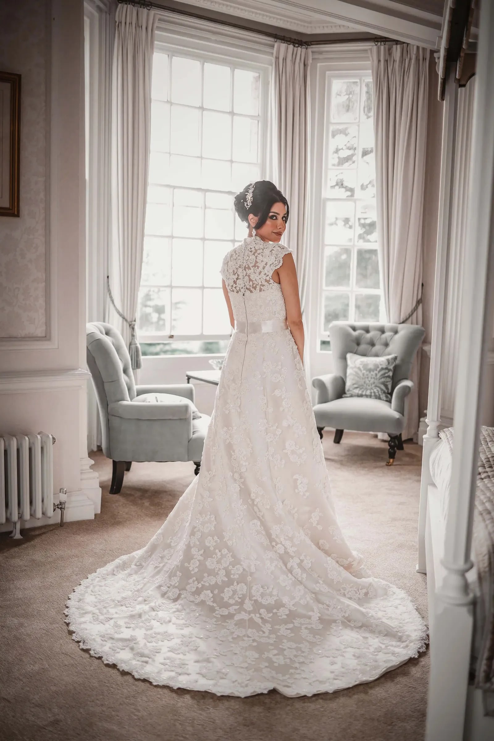 Bride looking back over her shoulder in a bay window, her lace train spread across the floor