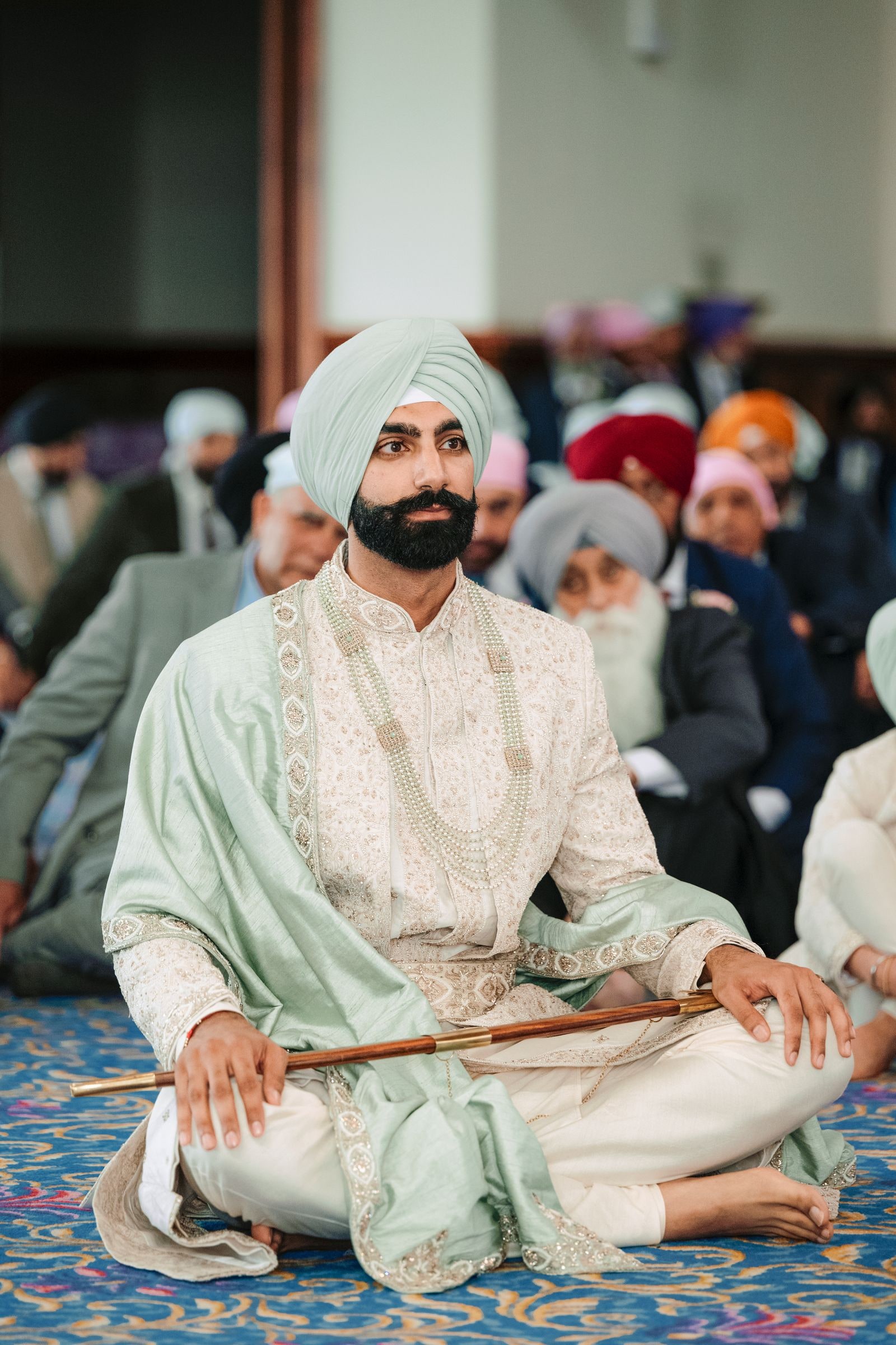 Groom seated cross-legged on the floor in a pale green turban, holding his kirpan