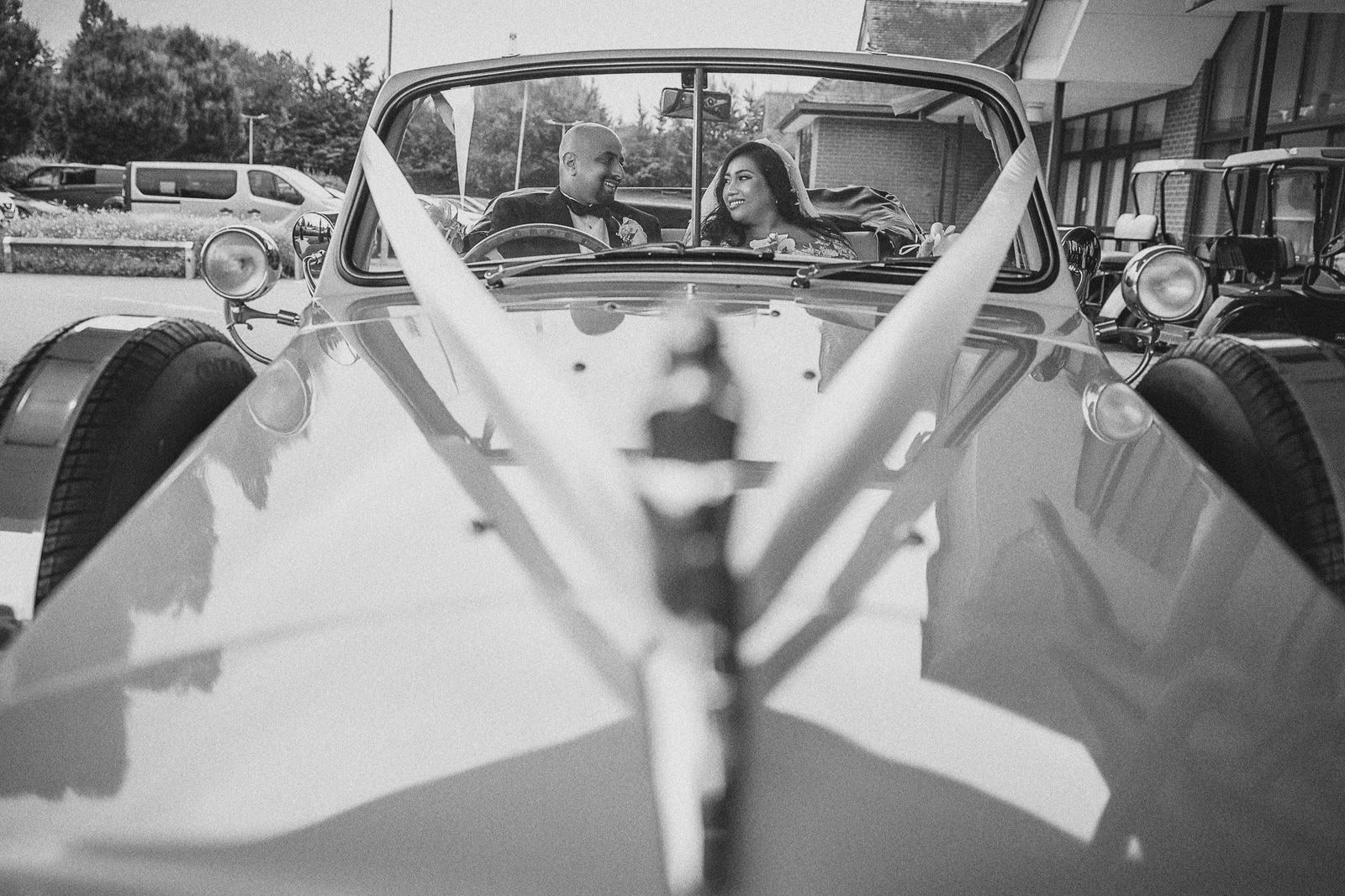 Bride and groom seated in an open-top vintage car dressed with ribbons, in black and white