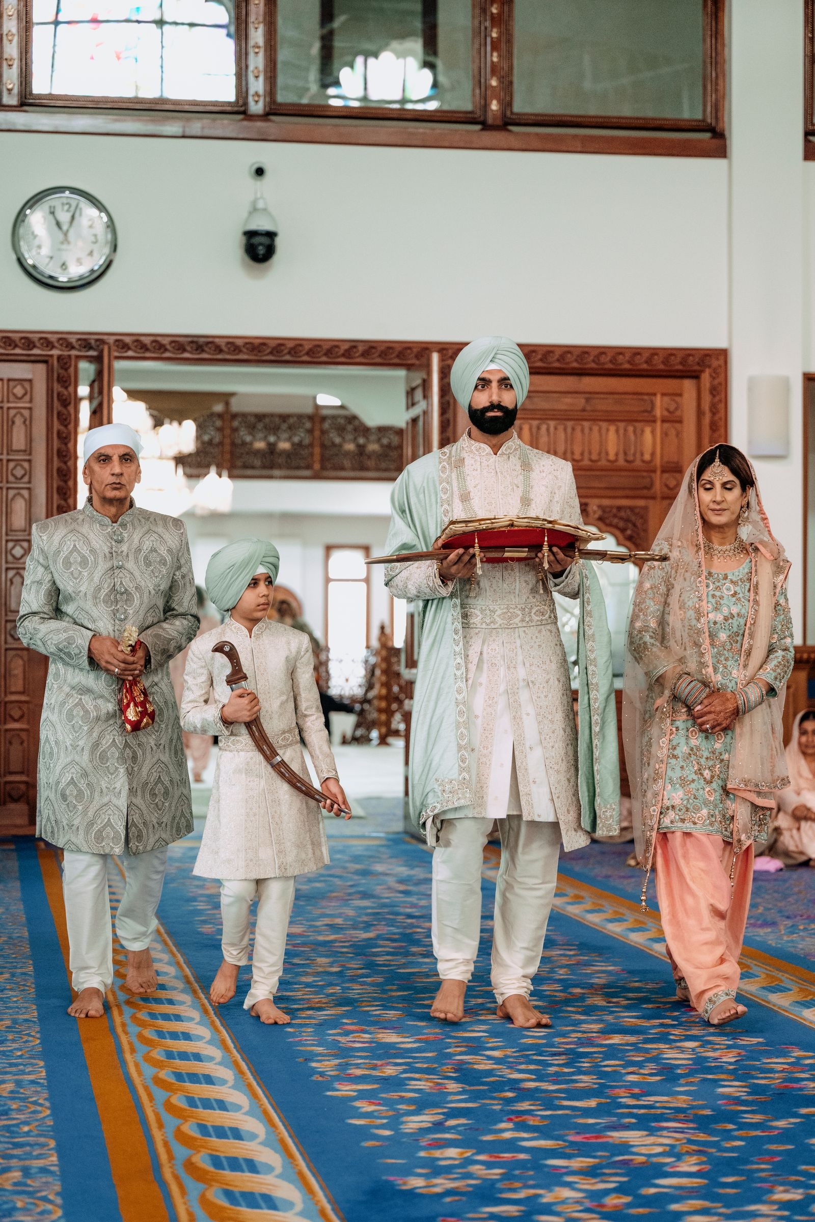 Groom carrying a tray up the aisle of the gurdwara, family walking with him