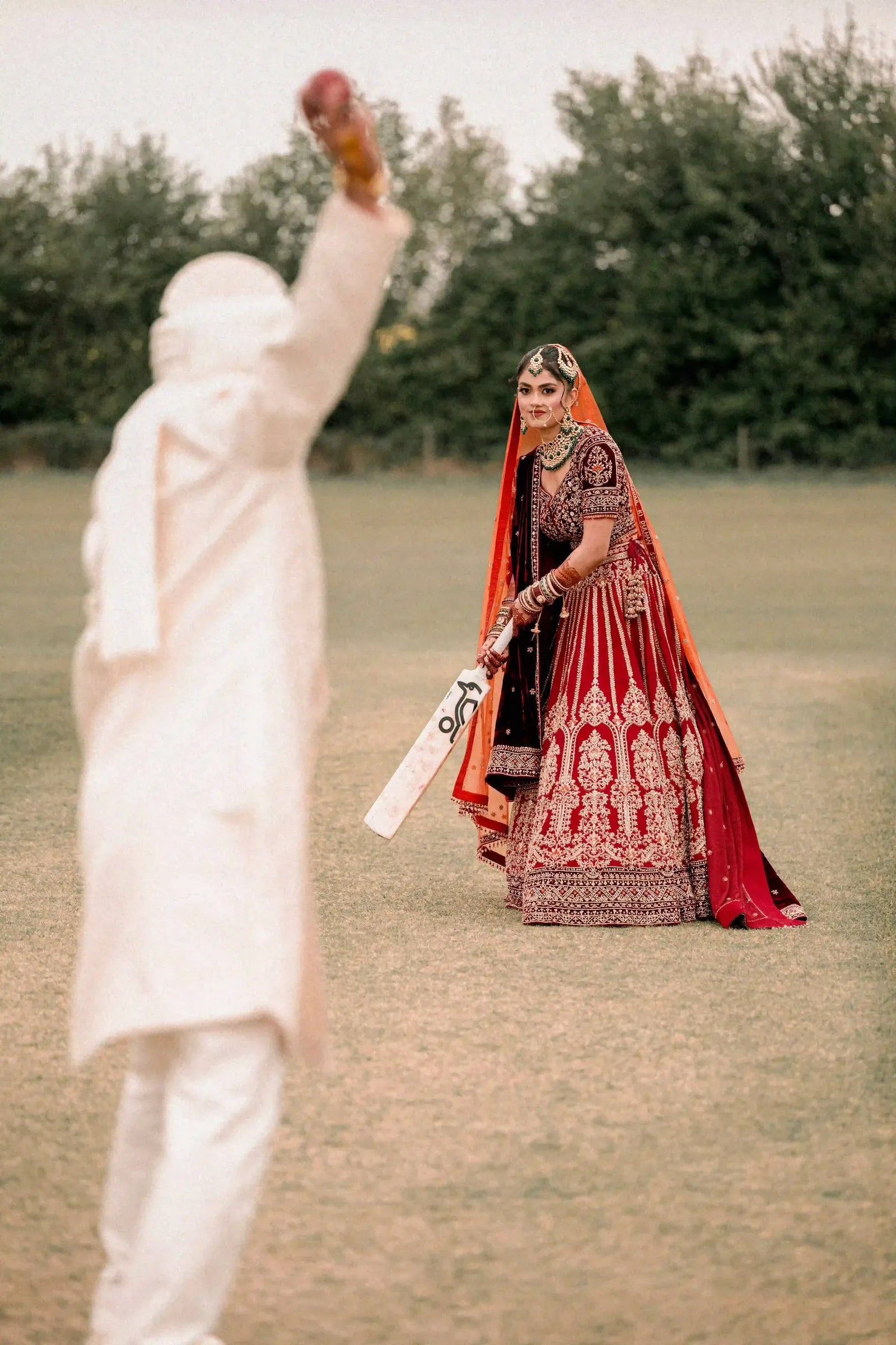Groom bowling a cricket ball to his bride, who is batting in her red lehenga