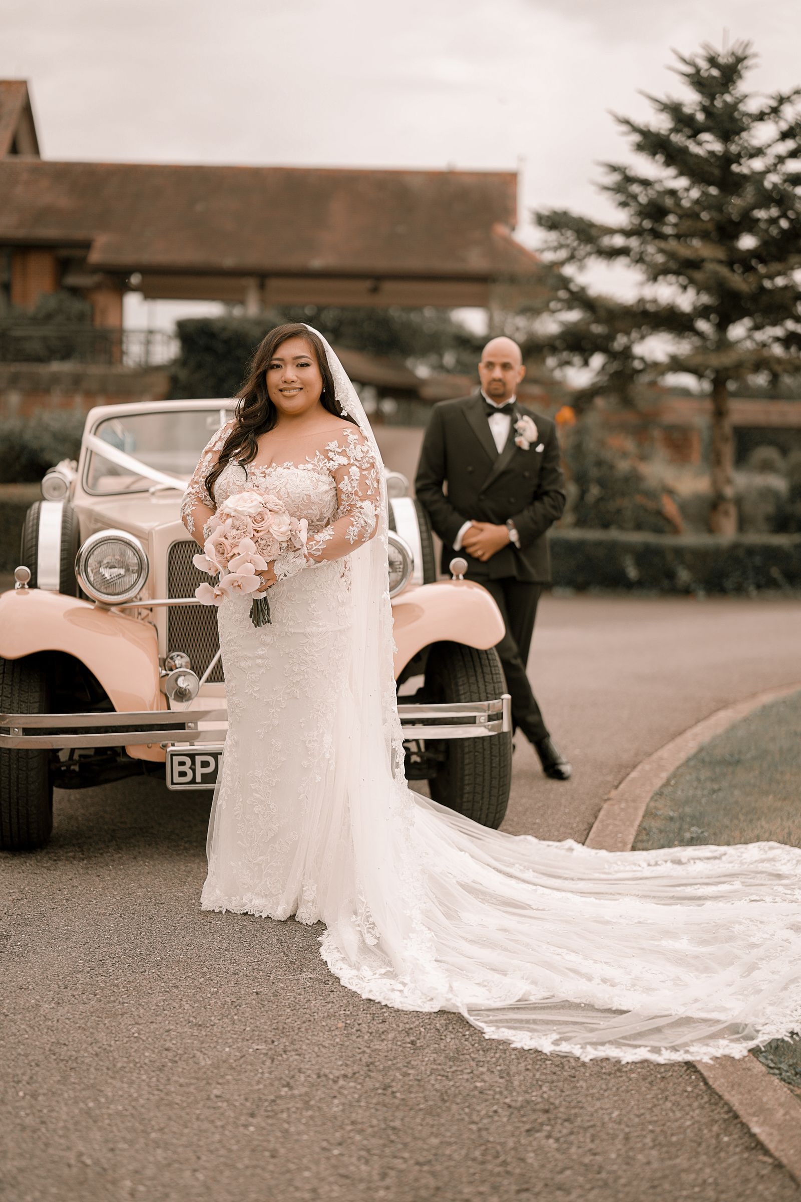 Bride beside the vintage car, her train spread across the tarmac, groom behind