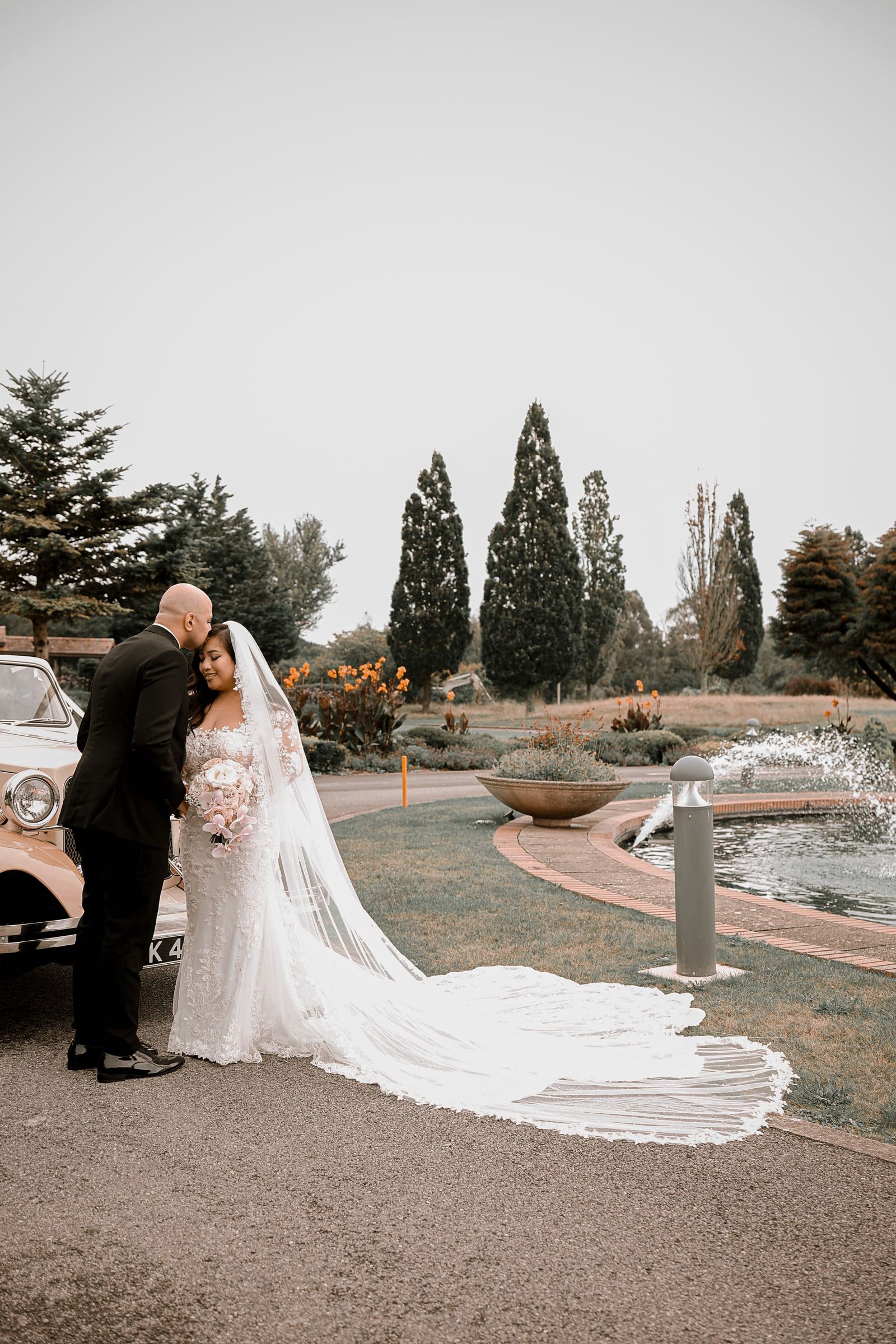 Groom kissing his bride's forehead by the car, her veil trailing on the ground