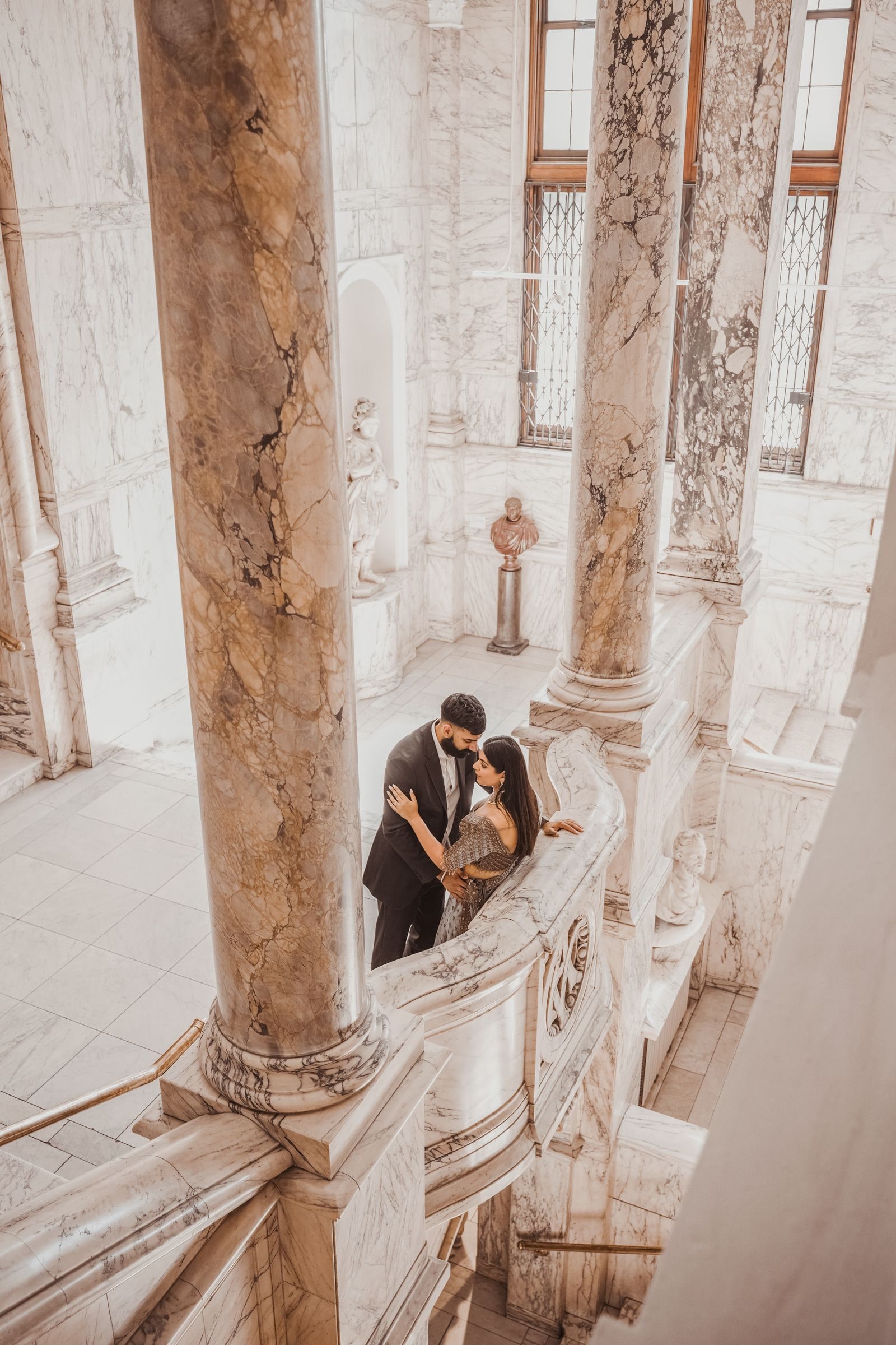 Couple small on a marble staircase seen from above, columns rising around them