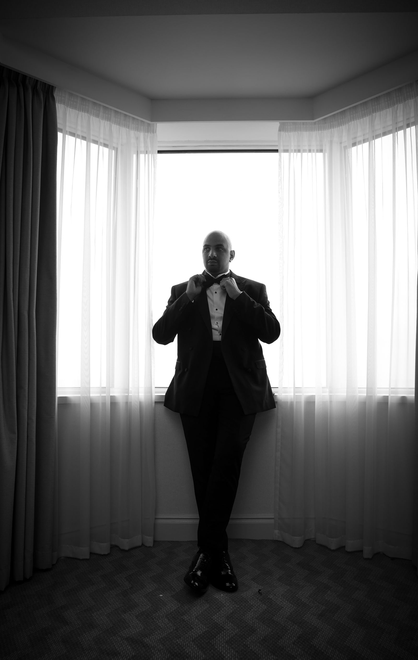 Groom fastening his bow tie in a bay window, in black and white