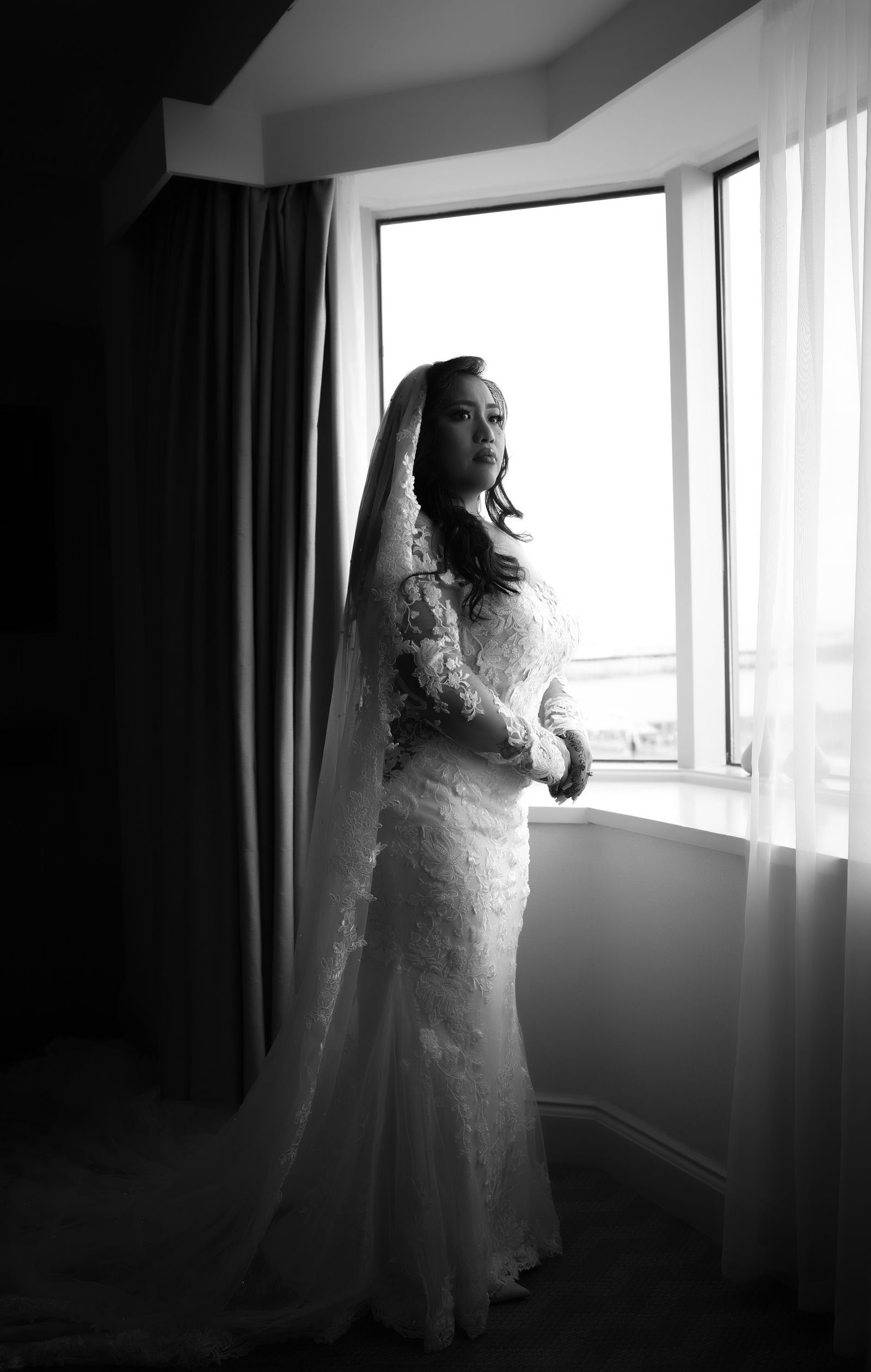 Bride standing veiled at a bright window, in black and white