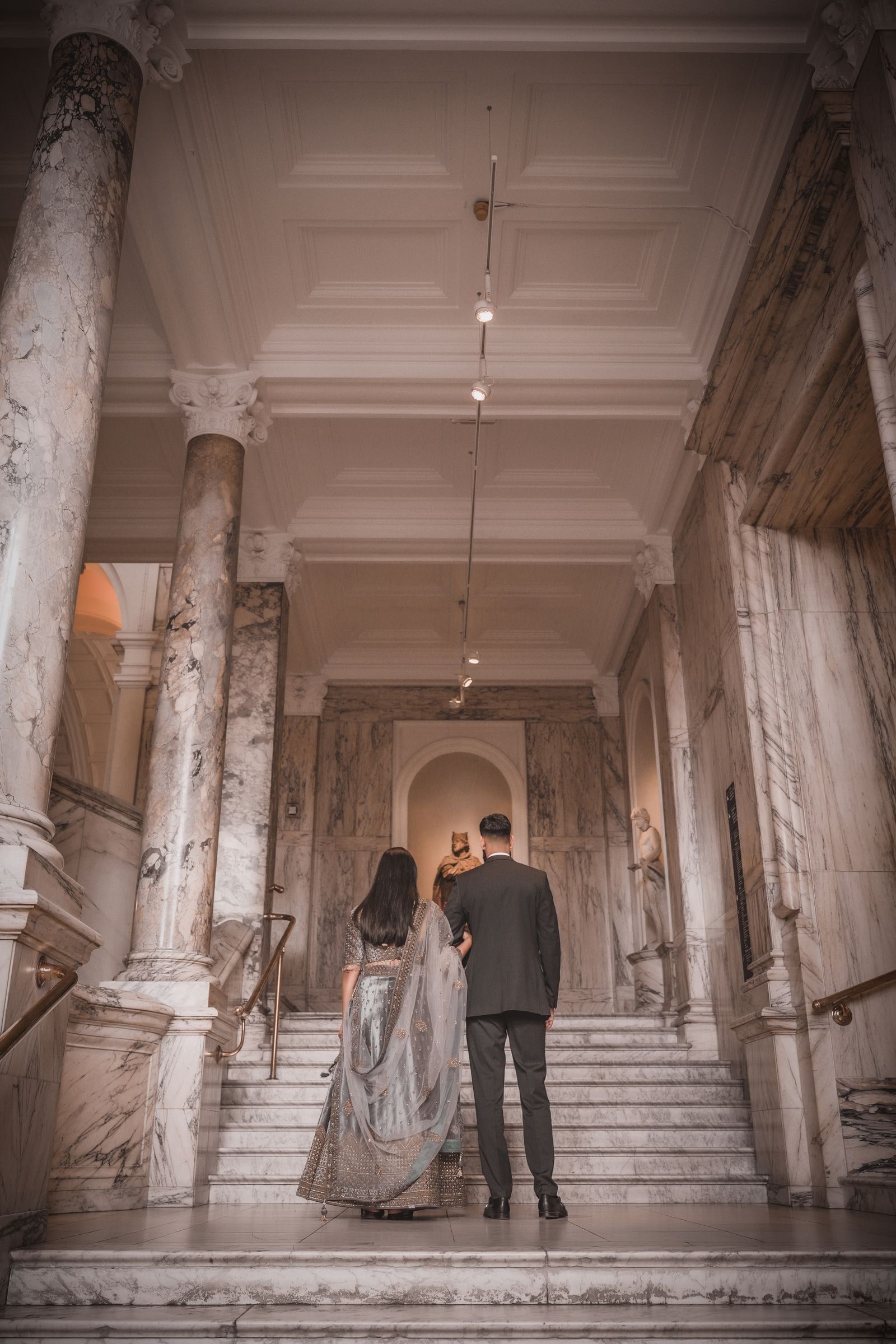 Couple seen from behind climbing the marble stairs down a long colonnade