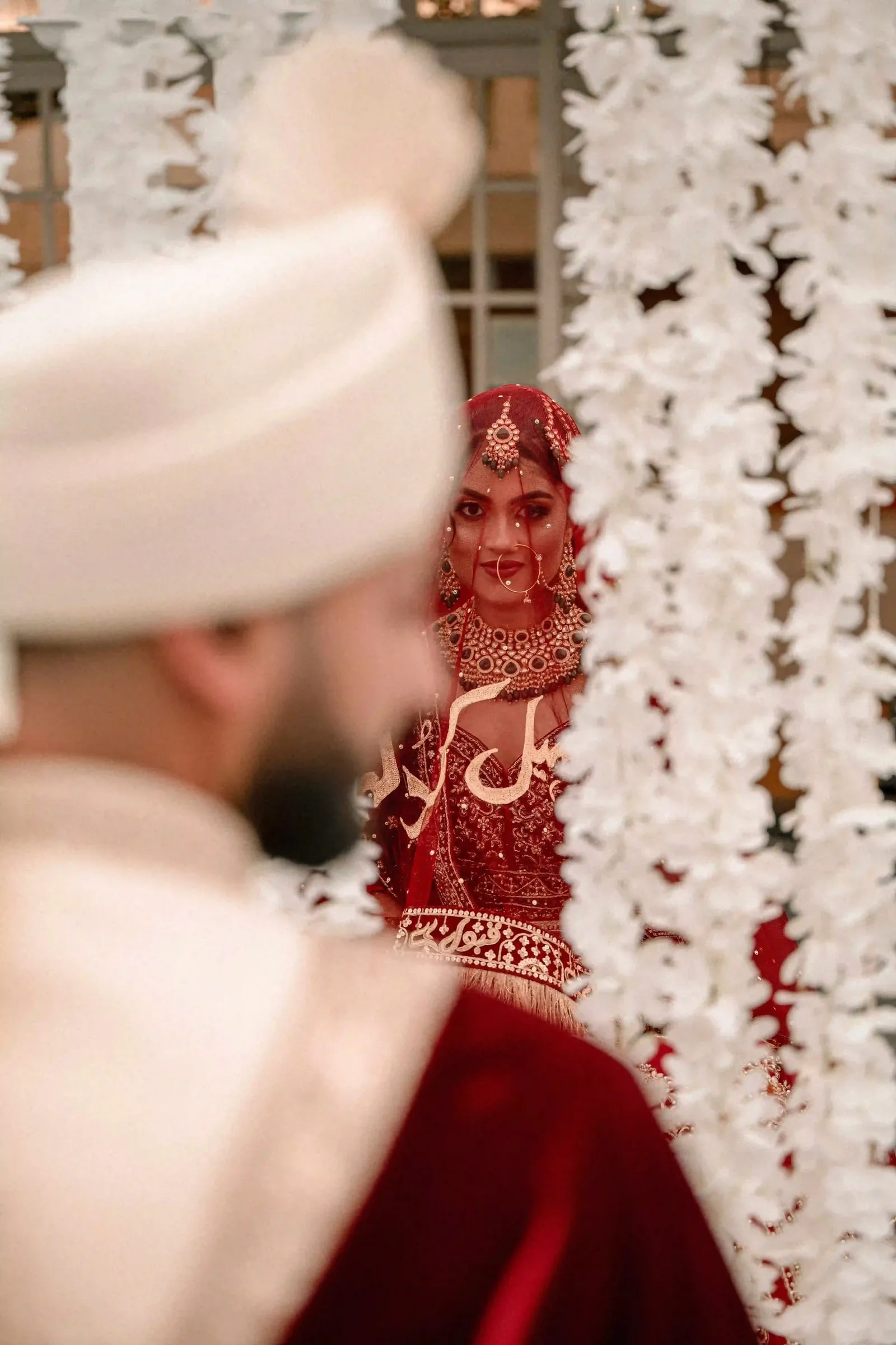 Bride framed past the groom's shoulder against a wall of white flowers