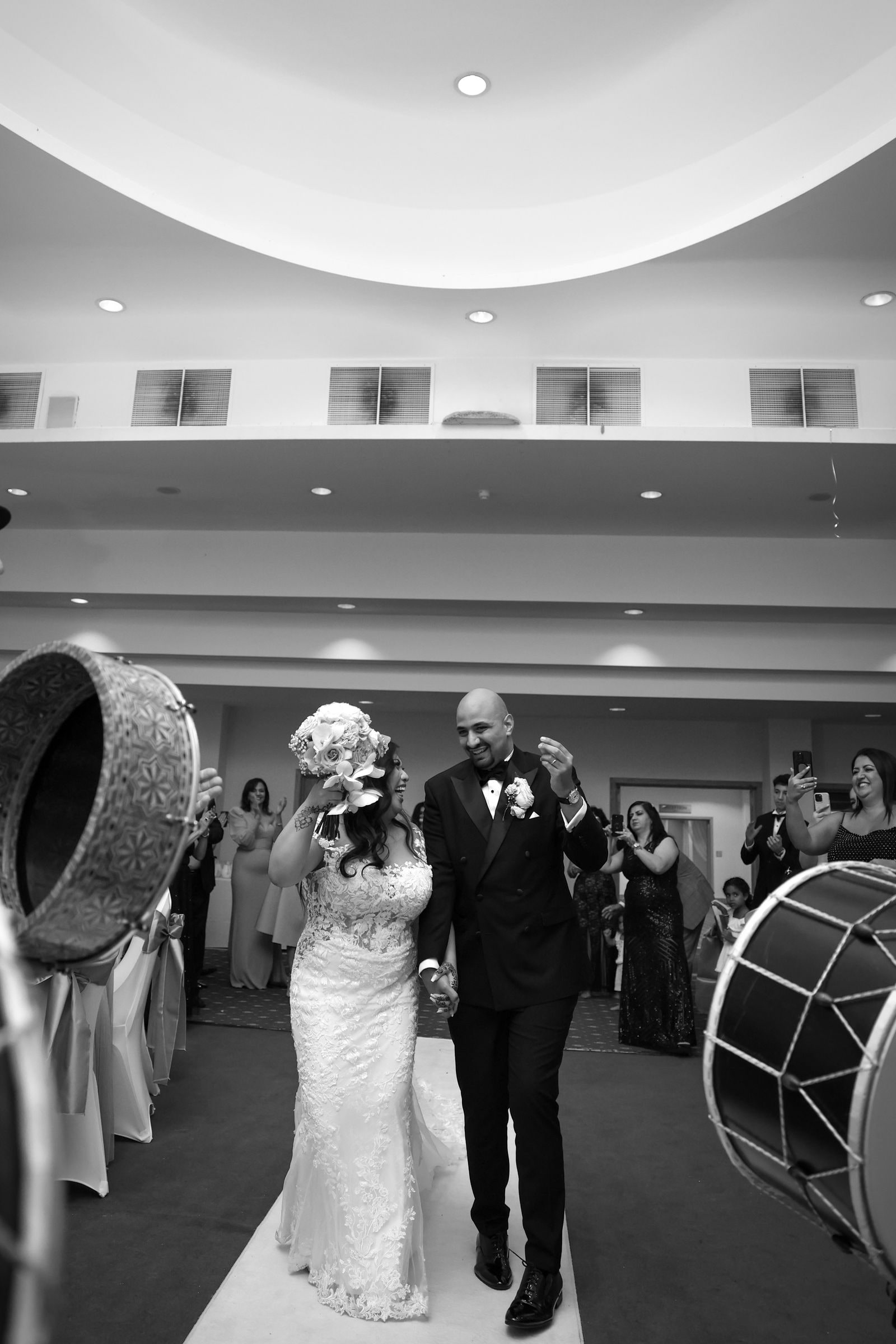 Bride and groom walking in past dhol players, guests applauding, in black and white