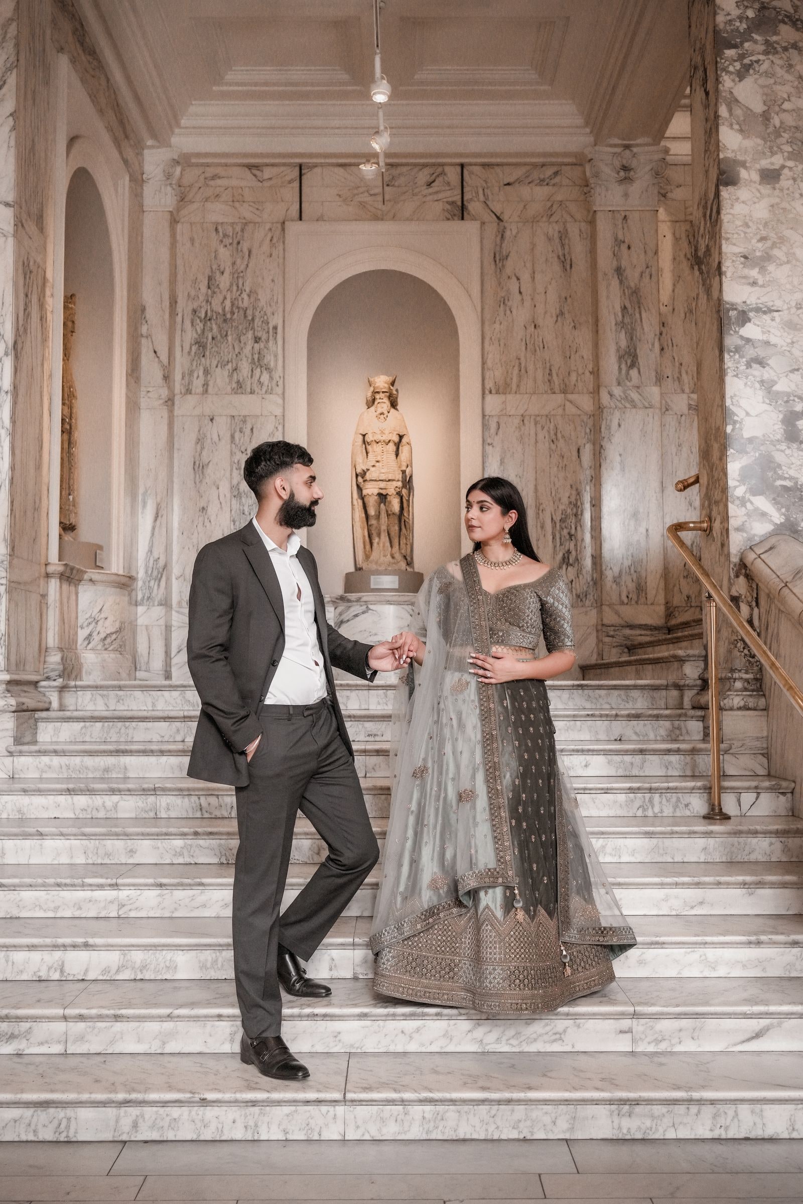 Bride to be and her fiance on the museum stairs, a gilded statue in the niche behind