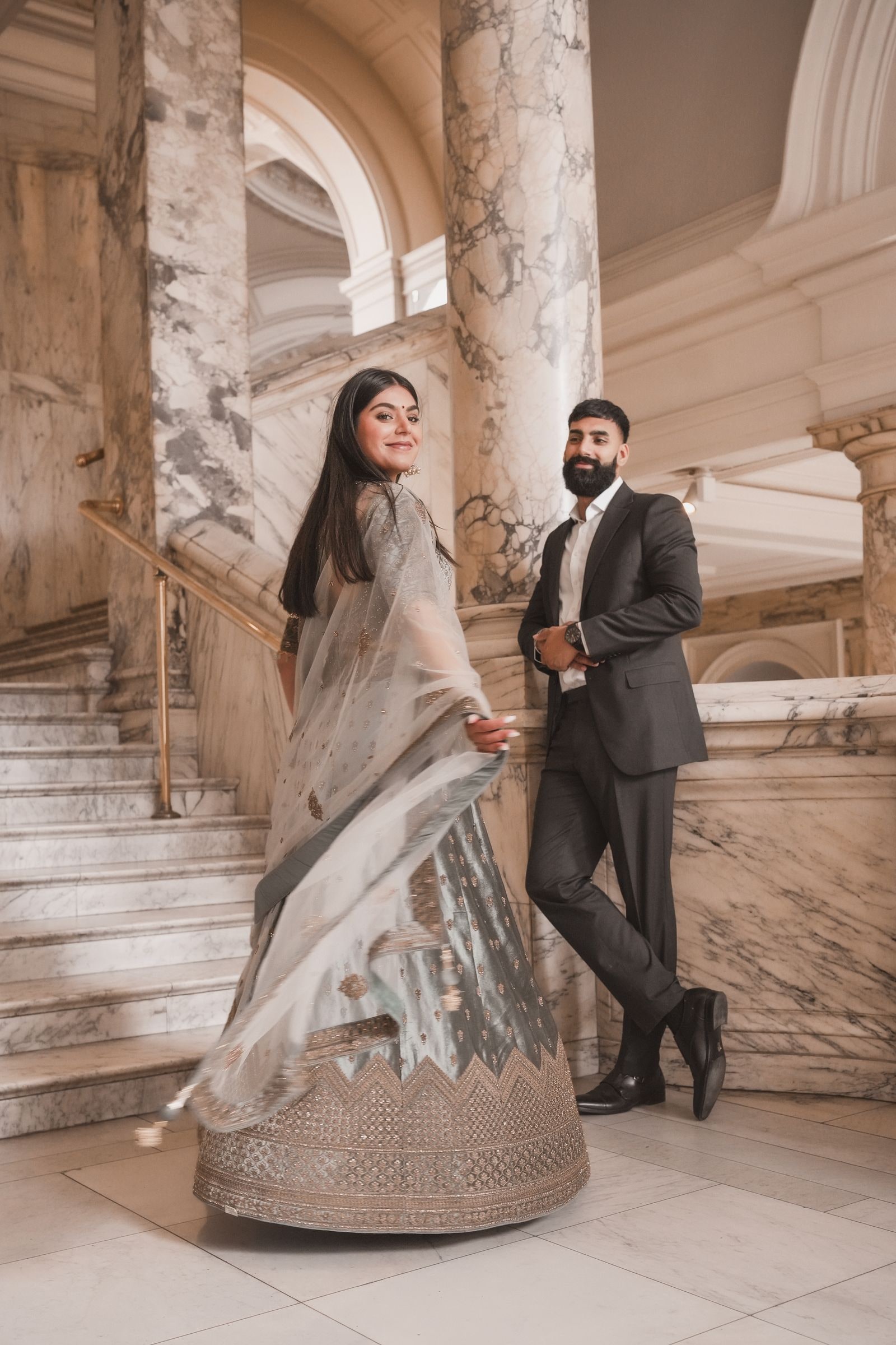 Couple standing on the marble staircase, her sage lehenga spread on the step