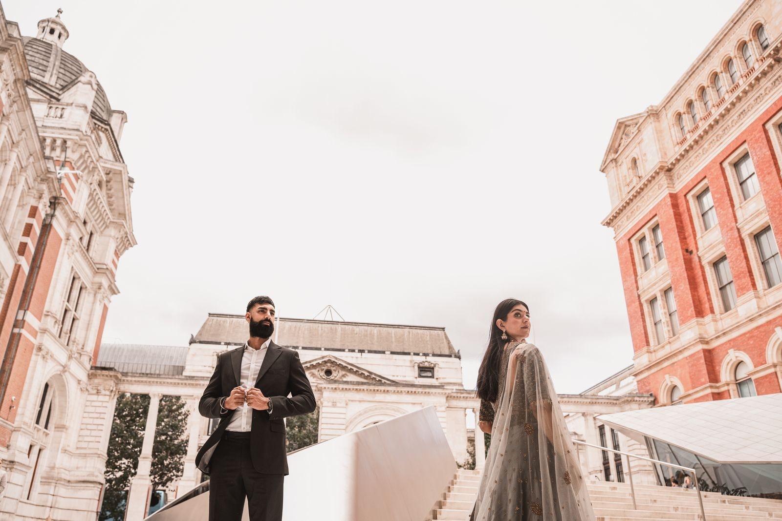 Couple standing apart on museum steps, red brick and stone buildings behind them