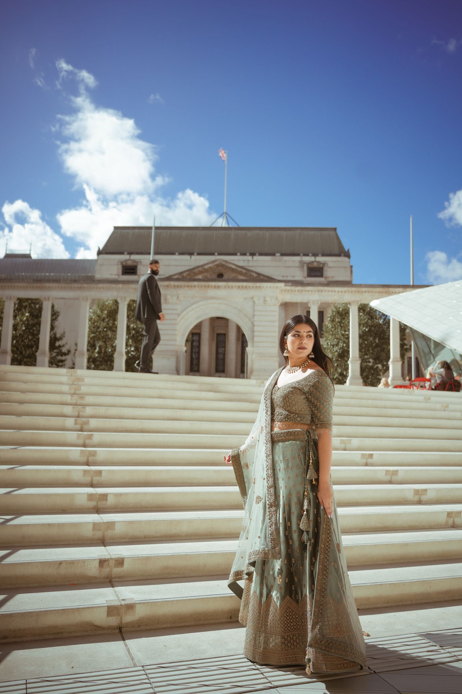 Woman in a sage and gold lehenga on wide stone steps under a blue sky