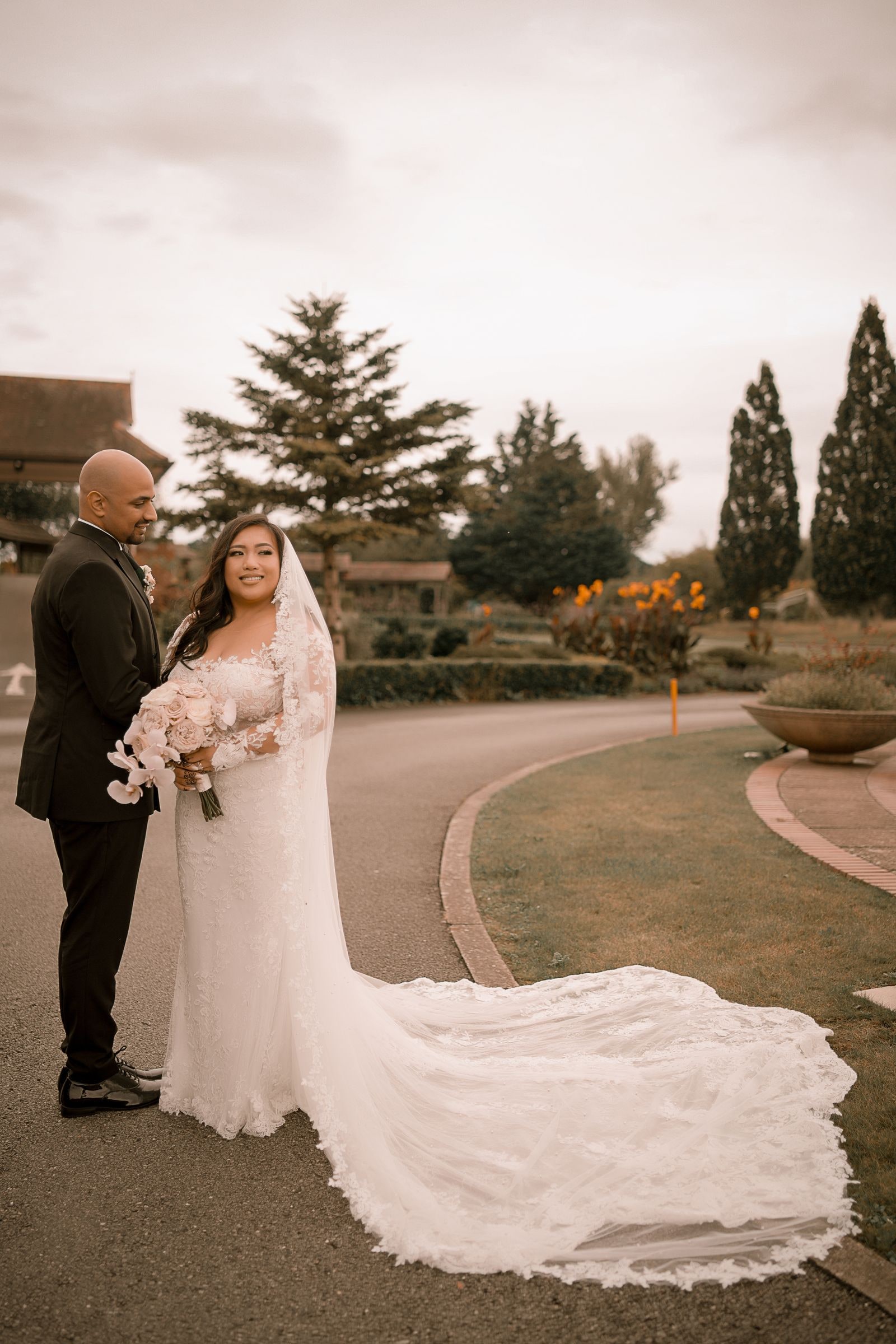 Bride in a lace gown with a long train and her groom in black tie on the drive