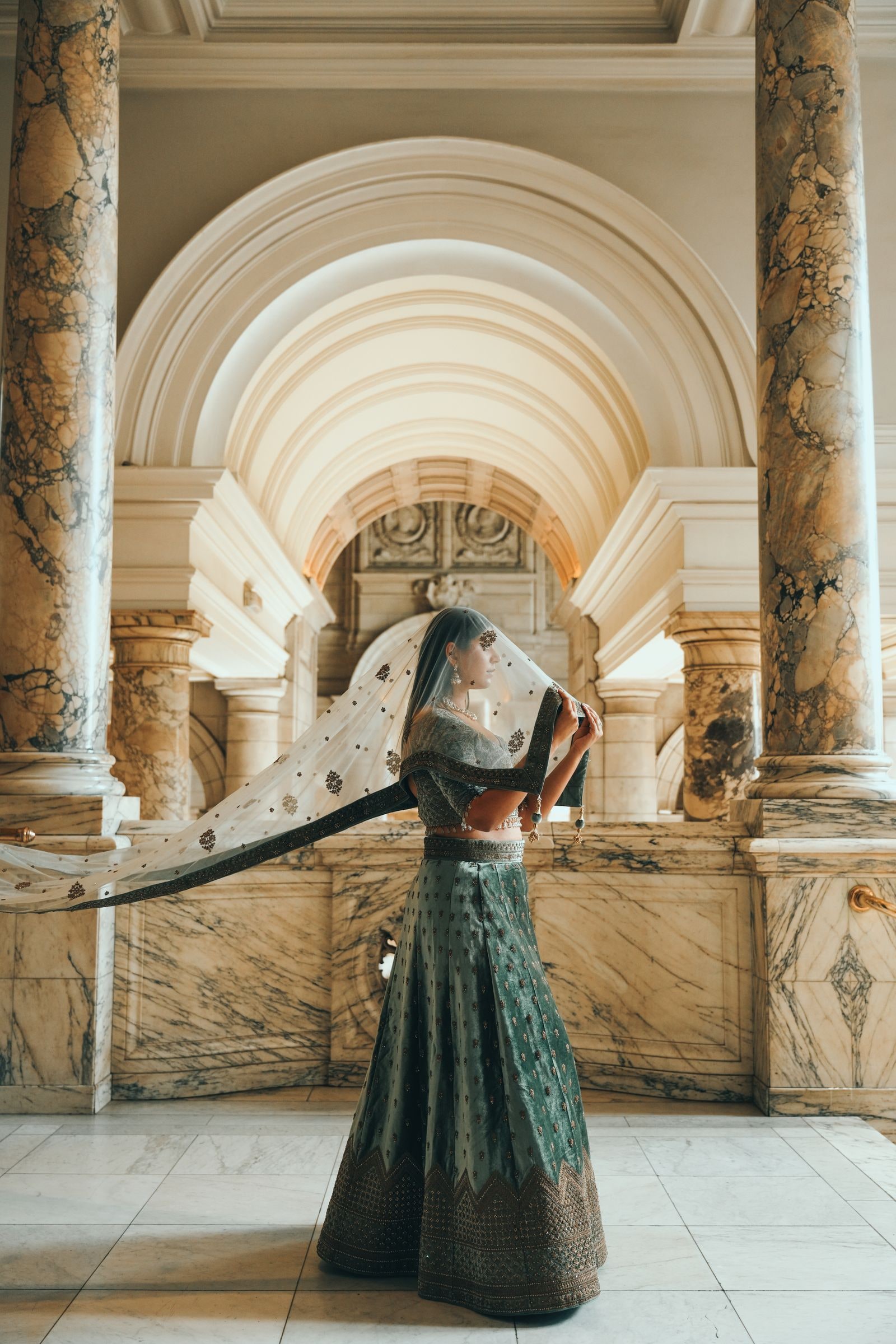 Woman turning in a dark green lehenga, her dupatta lifted, beneath a marble arch