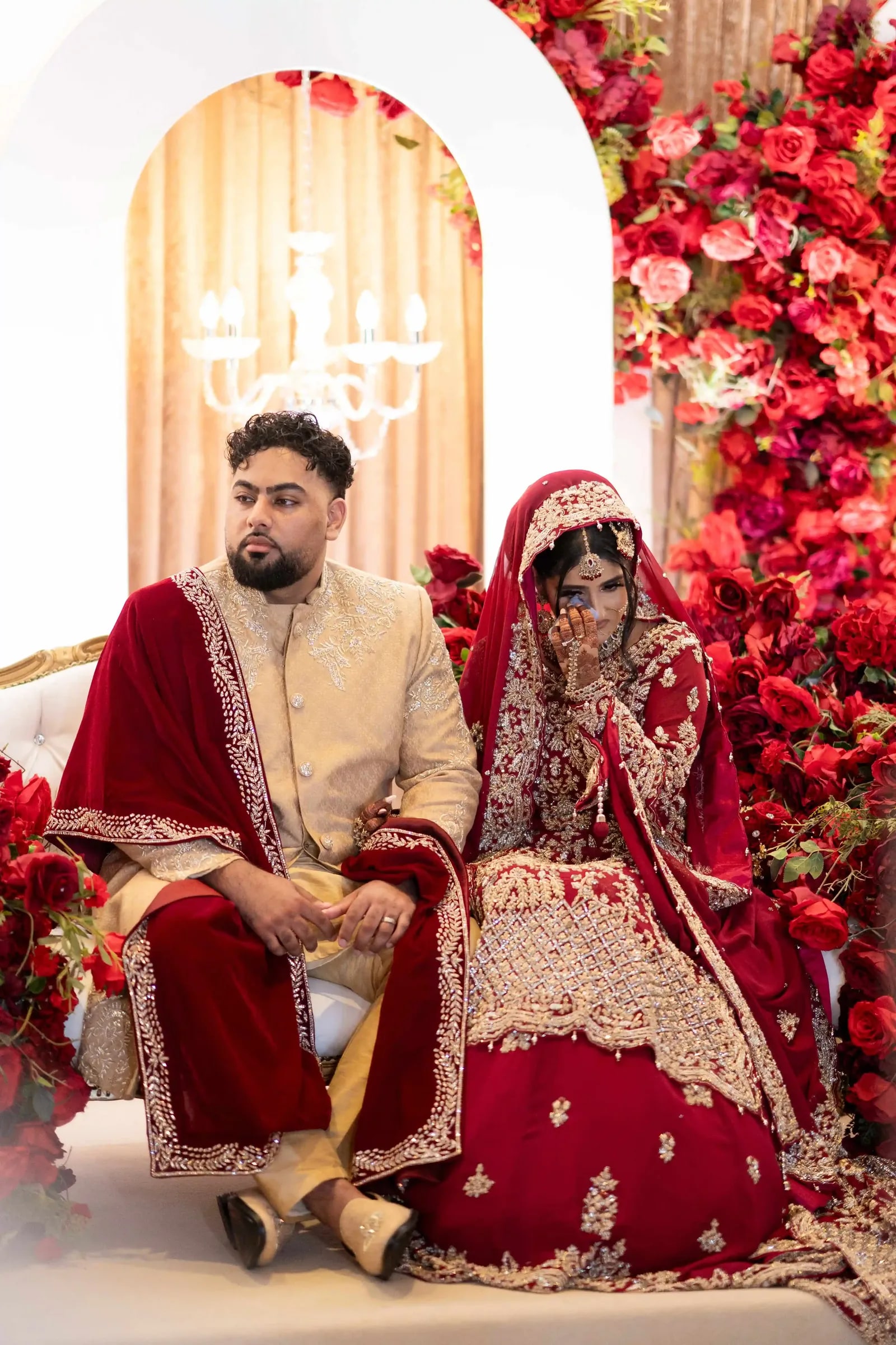 Bride and groom seated on the stage against a wall of red roses, he in a red velvet shawl